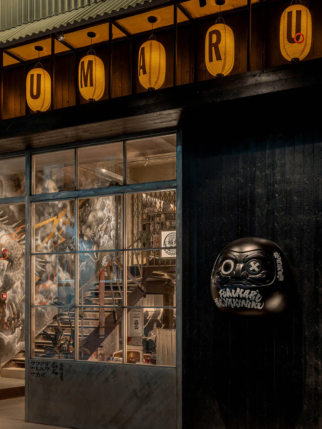 Night view of a Japanese yakiniku restaurant facade featuring large glass windows with dragon murals, hanging illuminated paper lanterns spelling UMARU, and a large black Daruma doll sign.