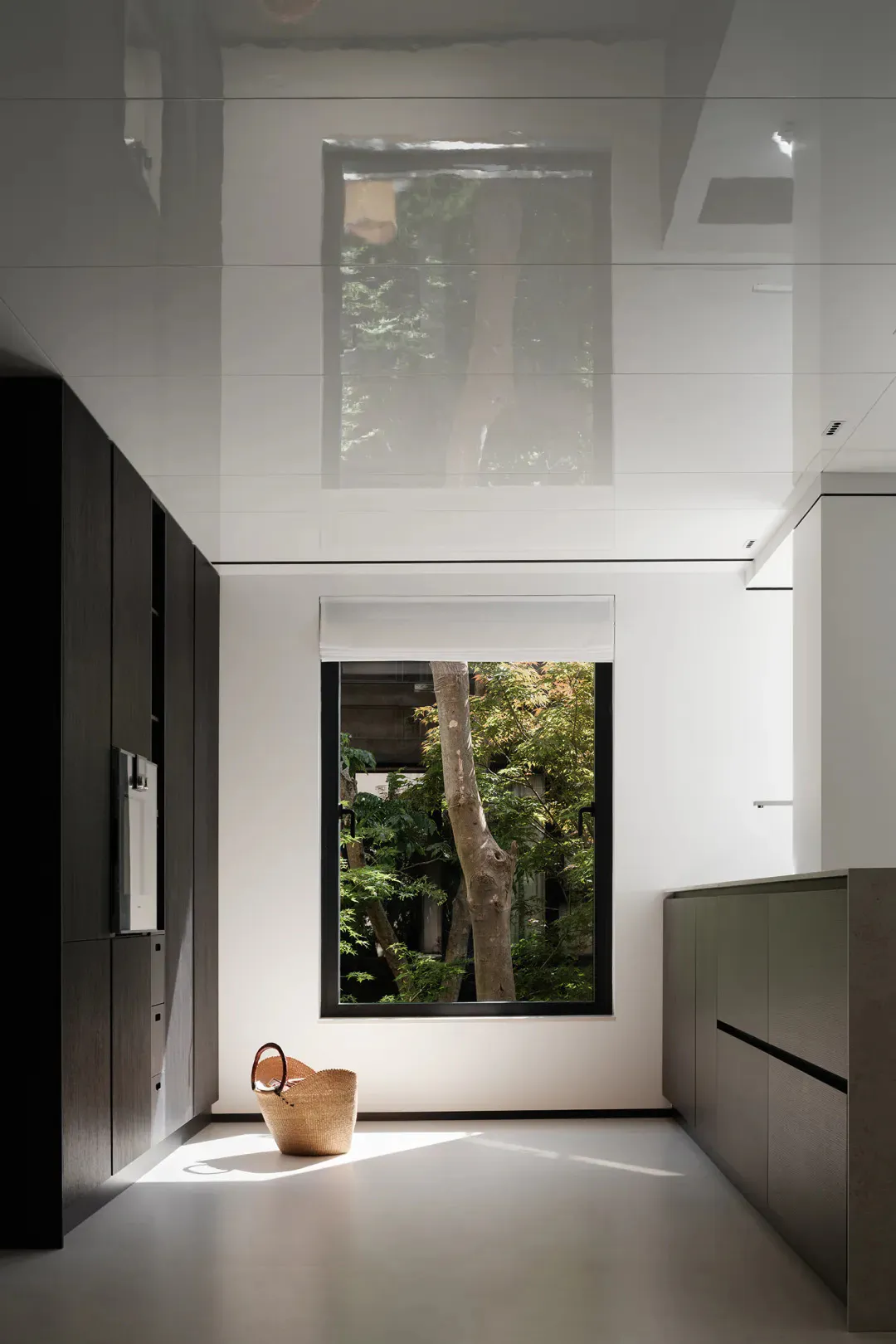 A modern minimalist kitchen with glossy white lacquered walls and cabinetry, a large vertical window showing green trees, a woven basket on the floor catching sunlight, and dark wood cabinetry on the left.