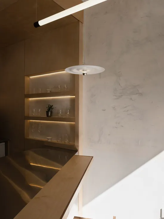 Modern minimalist bar area featuring light oak cabinetry, illuminated glass shelves, a round white pendant lamp, and a concrete wall.