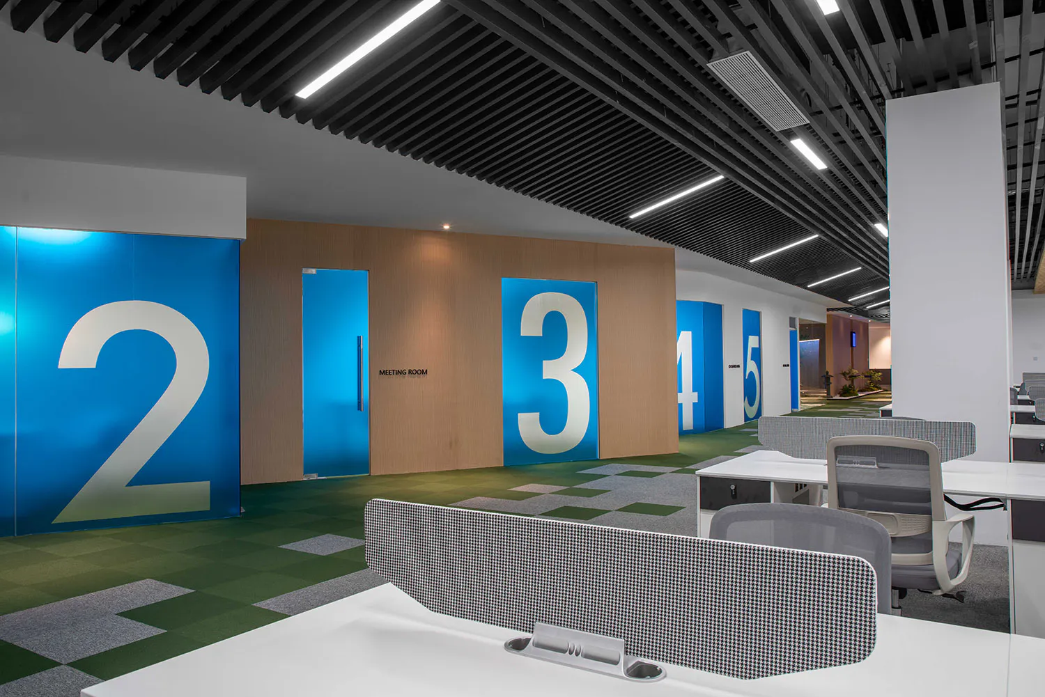 Modern office interior showing a hallway with large blue meeting room doors numbered 2, 3, 4, and 5, wood paneling, green and grey patterned carpet, white desks with ergonomic chairs in the foreground, and linear ceiling lighting.