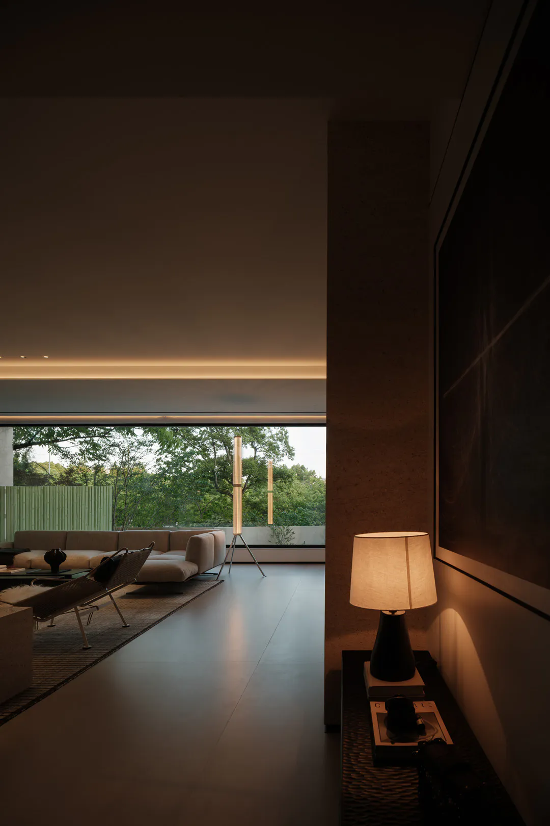 Modern minimalist living room featuring raw concrete walls, large floor-to-ceiling windows overlooking a green garden, a beige sectional sofa, a woven lounge chair, and warm ambient lighting from a tripod floor lamp and a table lamp.