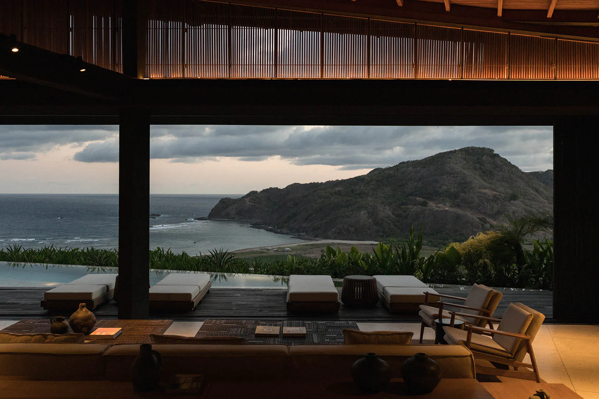 Interior view of a luxury villa's open living space with wooden ceiling slats, modern sofas and armchairs in the foreground, leading to an infinity pool deck with teak loungers and a sweeping view of the ocean, mountains, and dramatic cloudy sky at dusk.