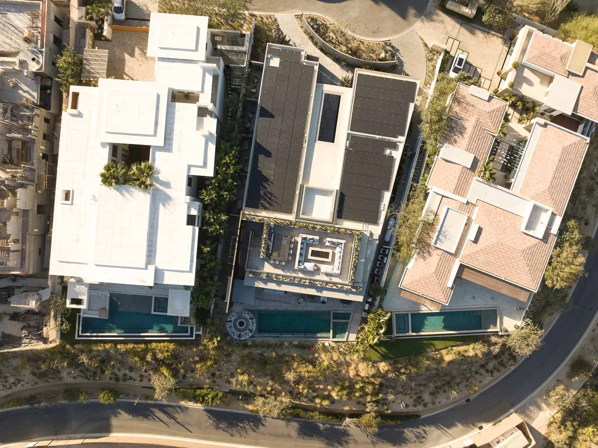 Aerial photograph of a modern luxury villa with white concrete architecture, dark solar panel roofs, rectangular swimming pools, and landscaped courtyards in a desert residential area.