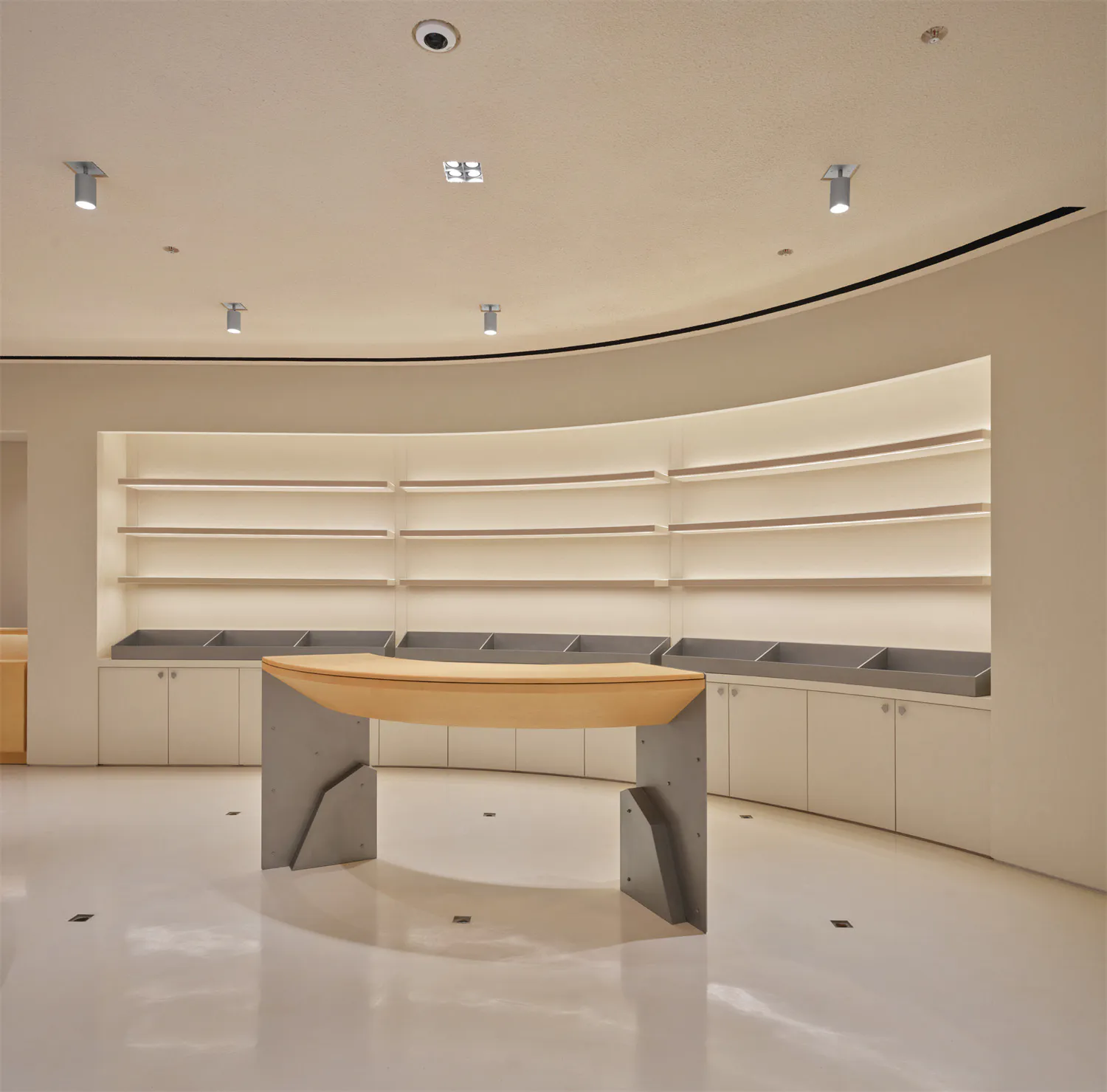 Modern minimalist retail interior featuring a curved light oak reception desk with angular concrete base, set against a curved wall with illuminated floating shelves and white glossy floor.