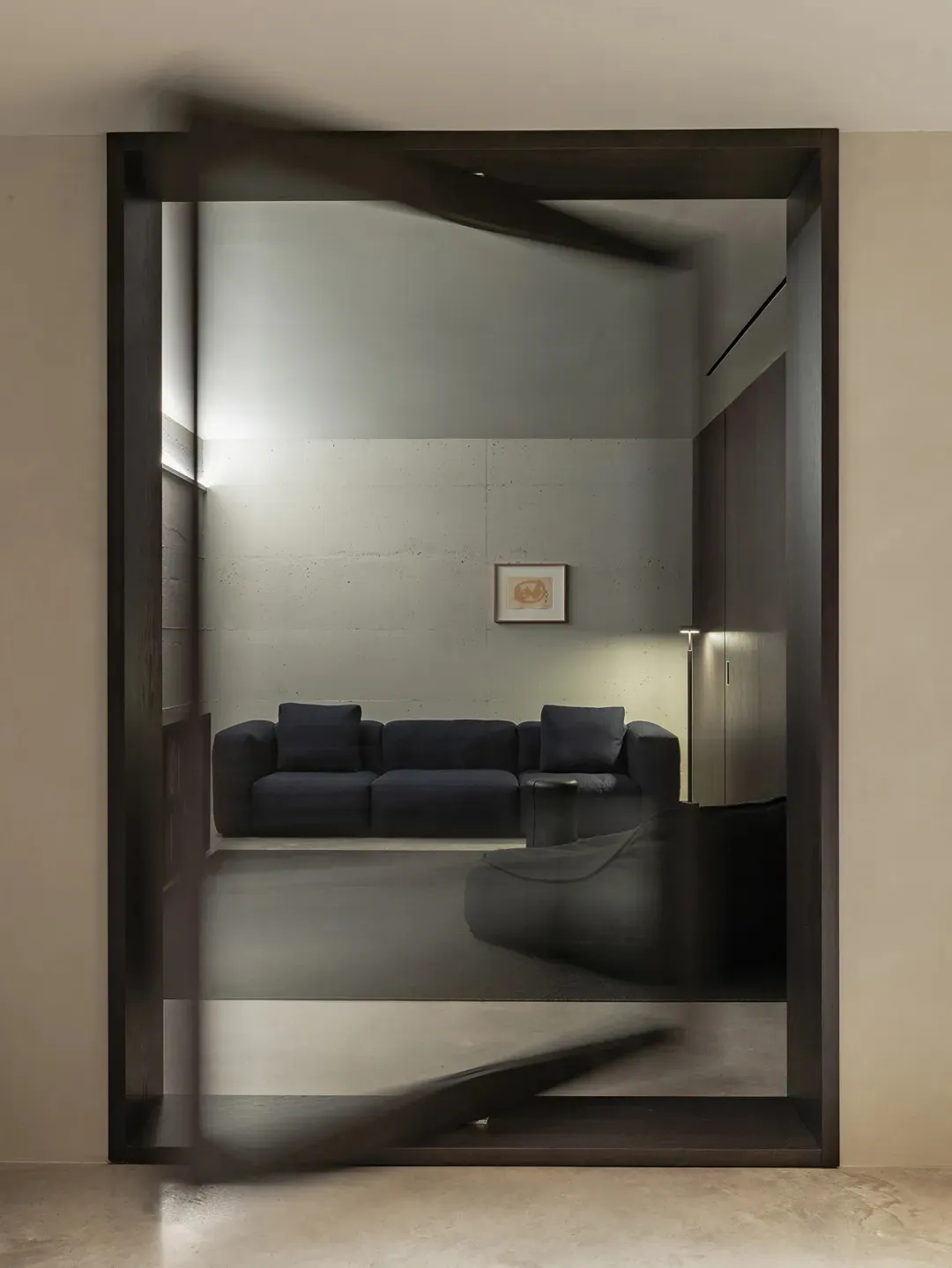 View through a large dark wooden pivot door into a minimalist living room featuring a concrete wall, dark blue sectional sofa, and modern lighting.