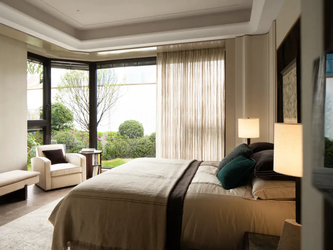 Modern serene bedroom interior with large bay window overlooking garden, neutral linen bedding, layered pillows in teal, dark brown and cream, two bedside lamps, and a reading armchair in the corner.