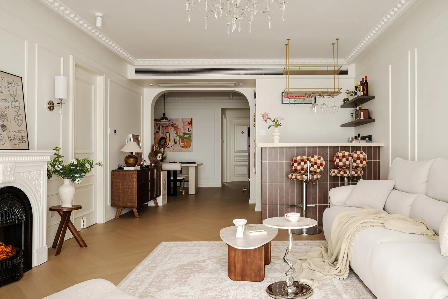 Bright and elegant living room with white sofa, marble coffee tables, white fireplace, and a brown checkerboard tiled bar area with bar stools in the background.