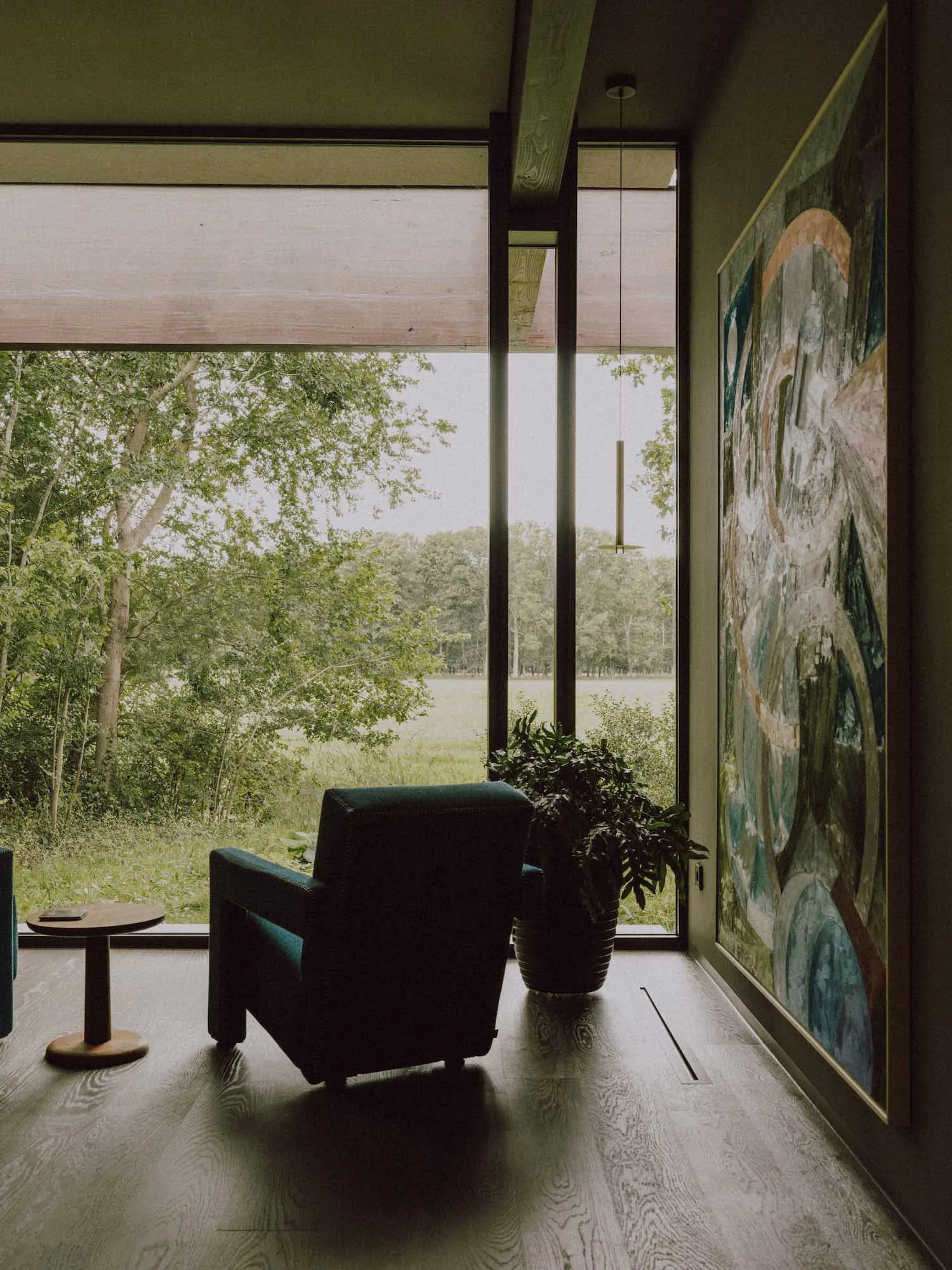Modern living room interior featuring a teal leather armchair, round wooden side table, large abstract painting, potted plant, and expansive floor-to-ceiling windows overlooking a green forest and meadow.