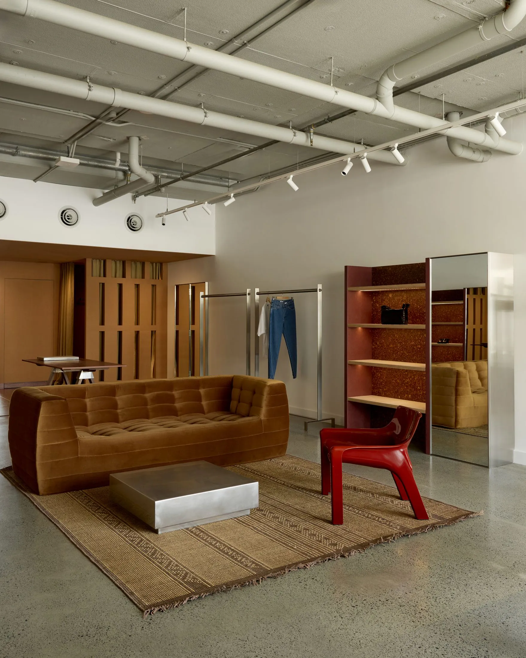 Industrial-style retail showroom with a brown tufted velvet sofa, metallic coffee table, red plastic chair, woven jute rug, and exposed white ceiling pipes.