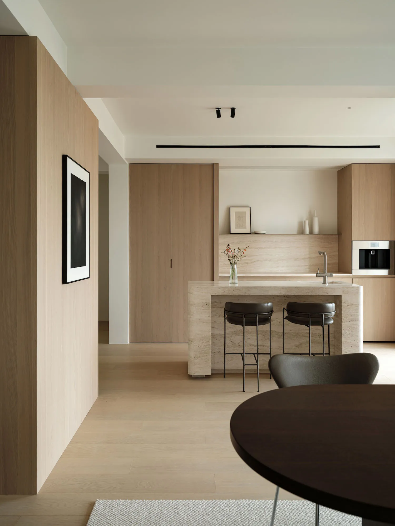 Modern minimalist kitchen with a large travertine stone island, two black leather bar stools, light oak wood cabinetry, and a round dark wood dining table in the foreground.