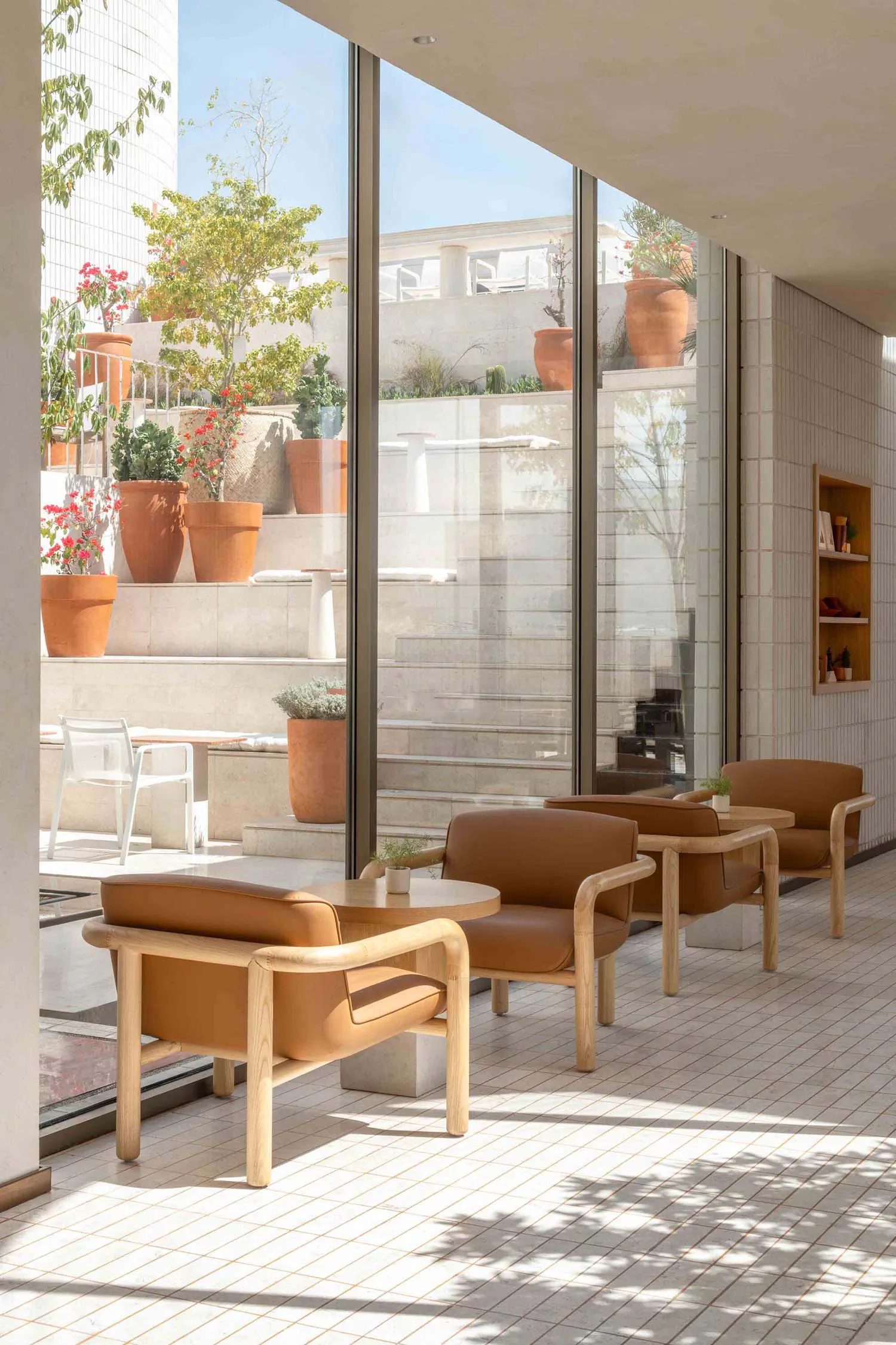 Modern indoor-outdoor lounge with three brown leather armchairs with light oak frames, round wooden tables, and a view of a sunny terrace filled with terracotta pots and flowering plants.
