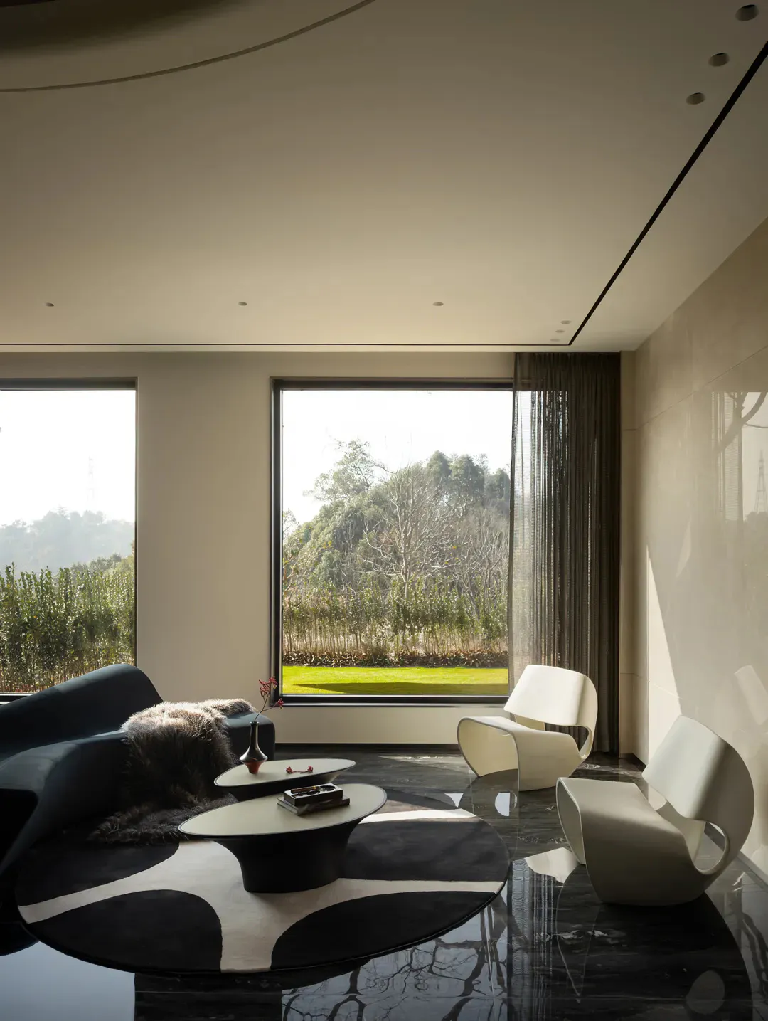 Modern minimalist living room featuring two white sculptural lounge chairs, a dark curved sofa with fur throw, black and white circular rug, and large floor-to-ceiling windows overlooking a green garden and trees.