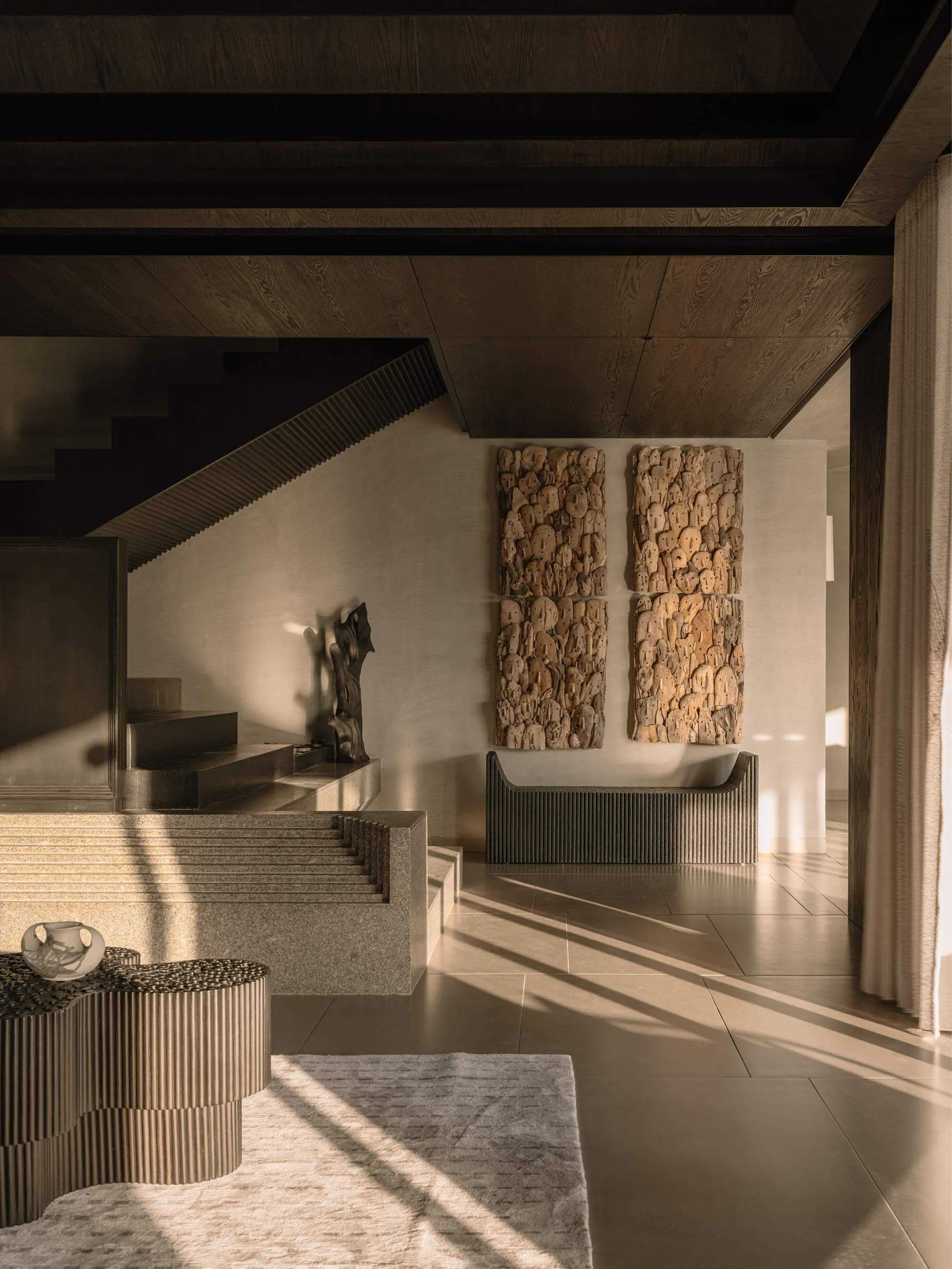 Modern minimalist entryway featuring travertine stairs, fluted stone furniture, two large organic stone wall sculptures, and a dark wood sculpture under dramatic natural light.