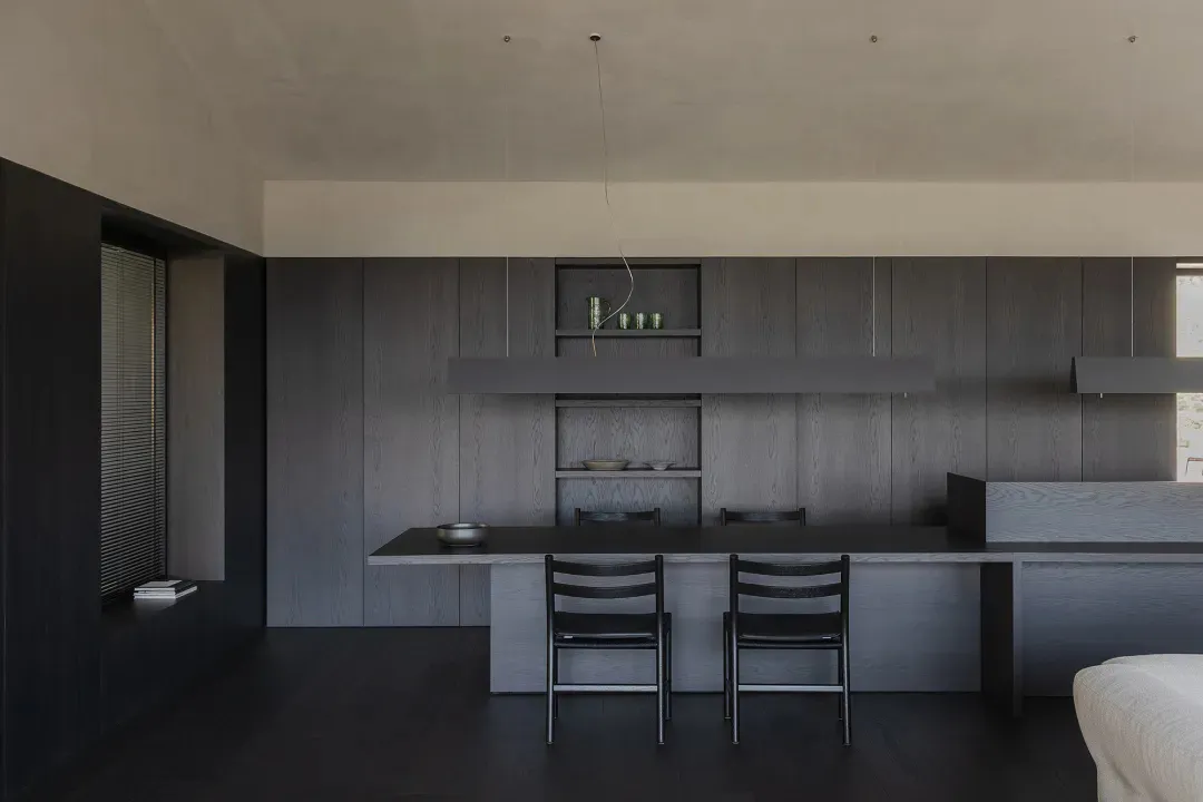 Modern minimalist kitchen featuring a long dark wood dining island with two black chairs, matching dark wood cabinetry, open shelving with ceramics, and a concrete ceiling with pendant lights.