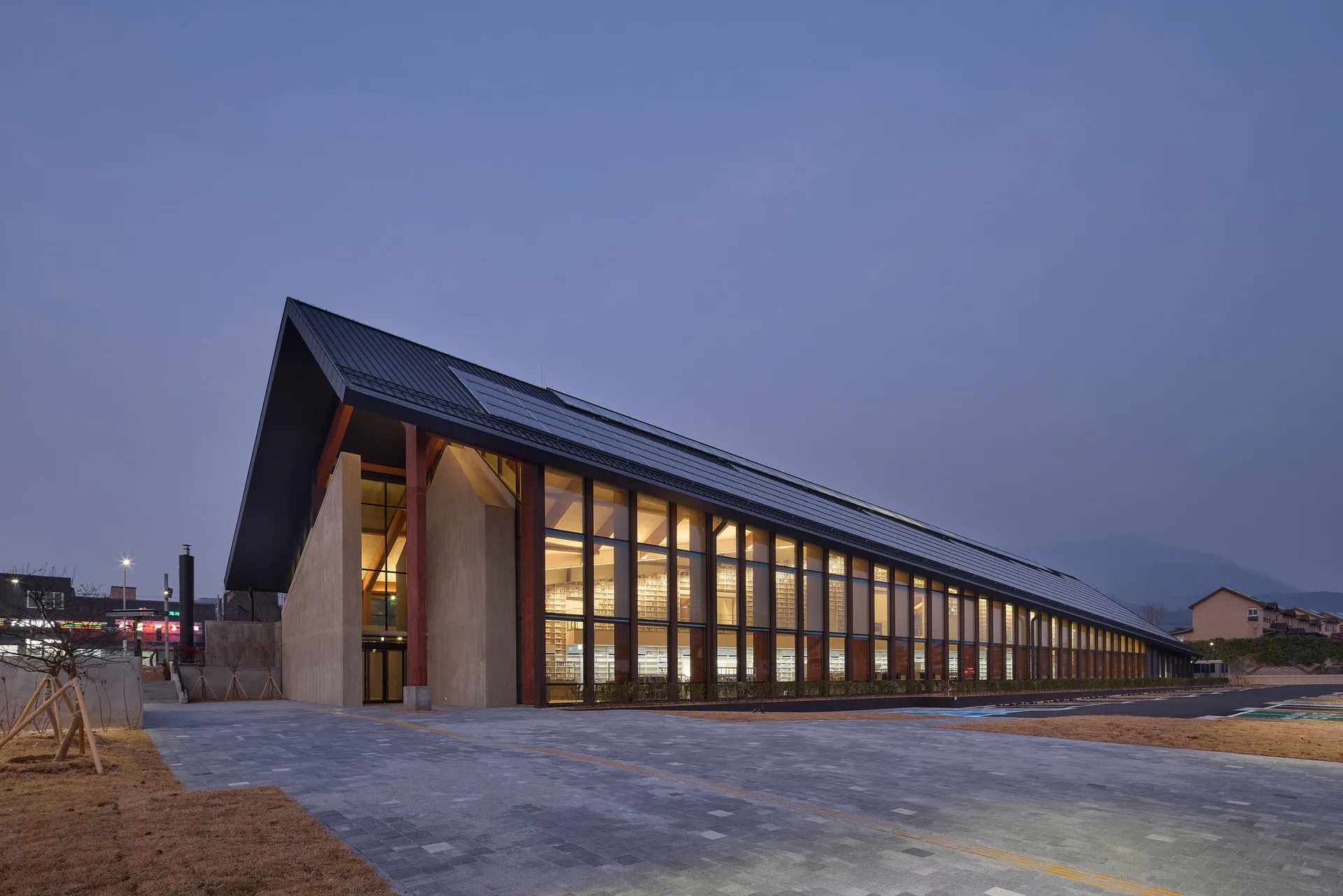 Exterior view of a long modern public library building at dusk, featuring a dramatic pitched roof, extensive floor-to-ceiling glass walls revealing glowing bookshelves inside, concrete and wood structural elements, set against mountains in Taiwan.
