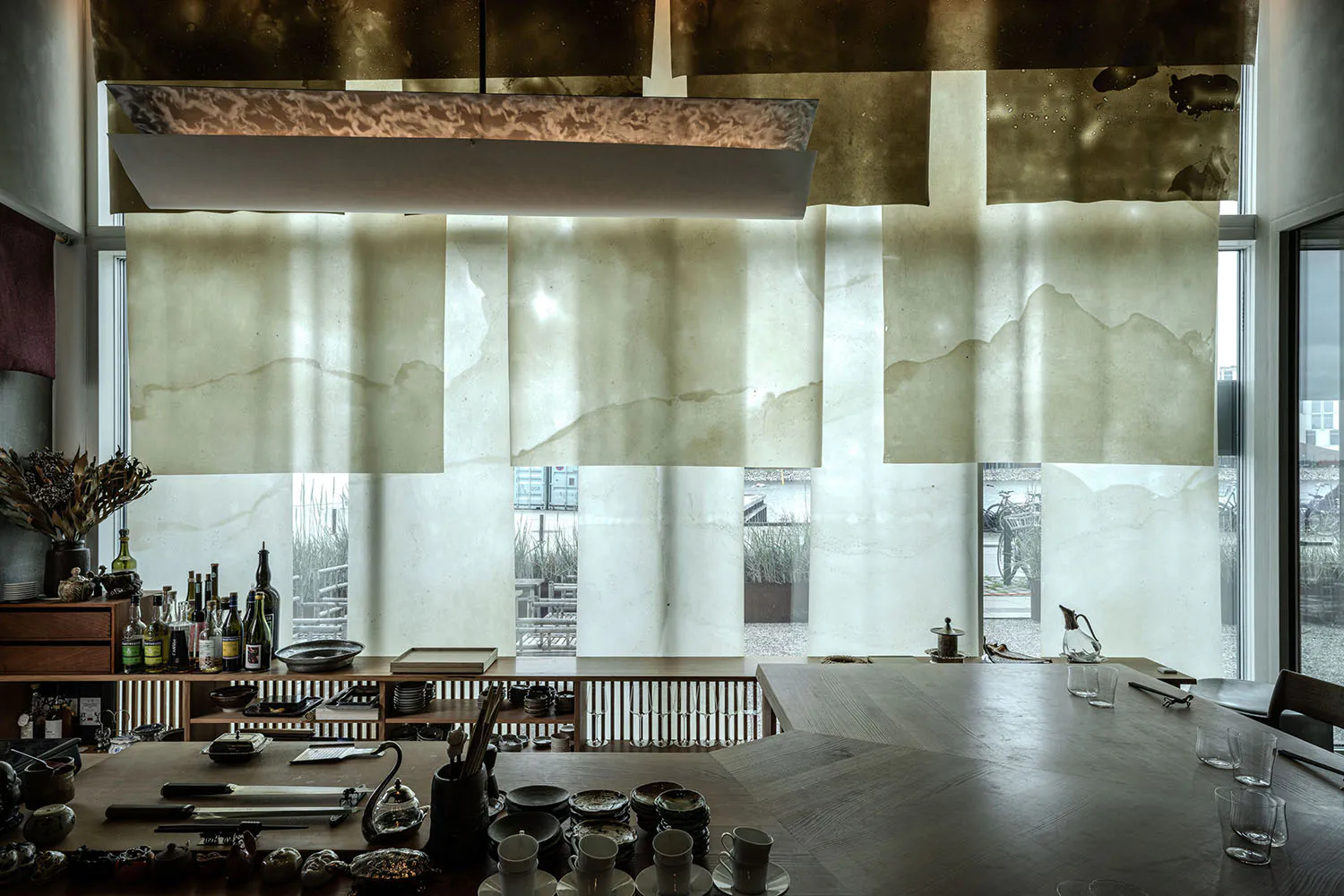 Modern dining room interior featuring a large rectangular patina brass pendant light, wooden dining table set with ceramics and glassware, and large windows with translucent shades overlooking a harbor with bicycles and water views.