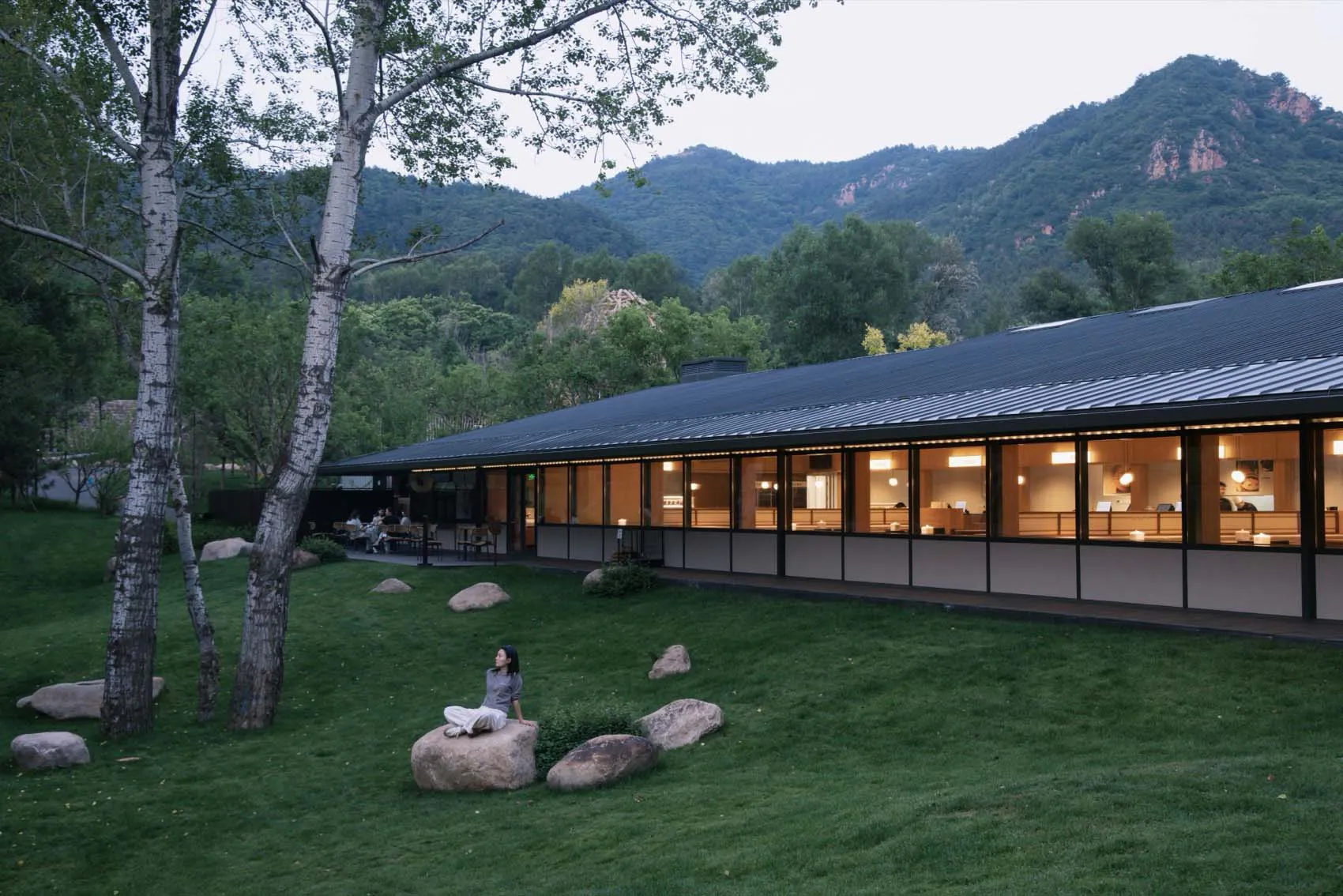 A contemporary mountain lodge with extensive glass windows glowing warmly at dusk, set against forested mountains, with a woman in white sitting on a large rock in the foreground lawn.