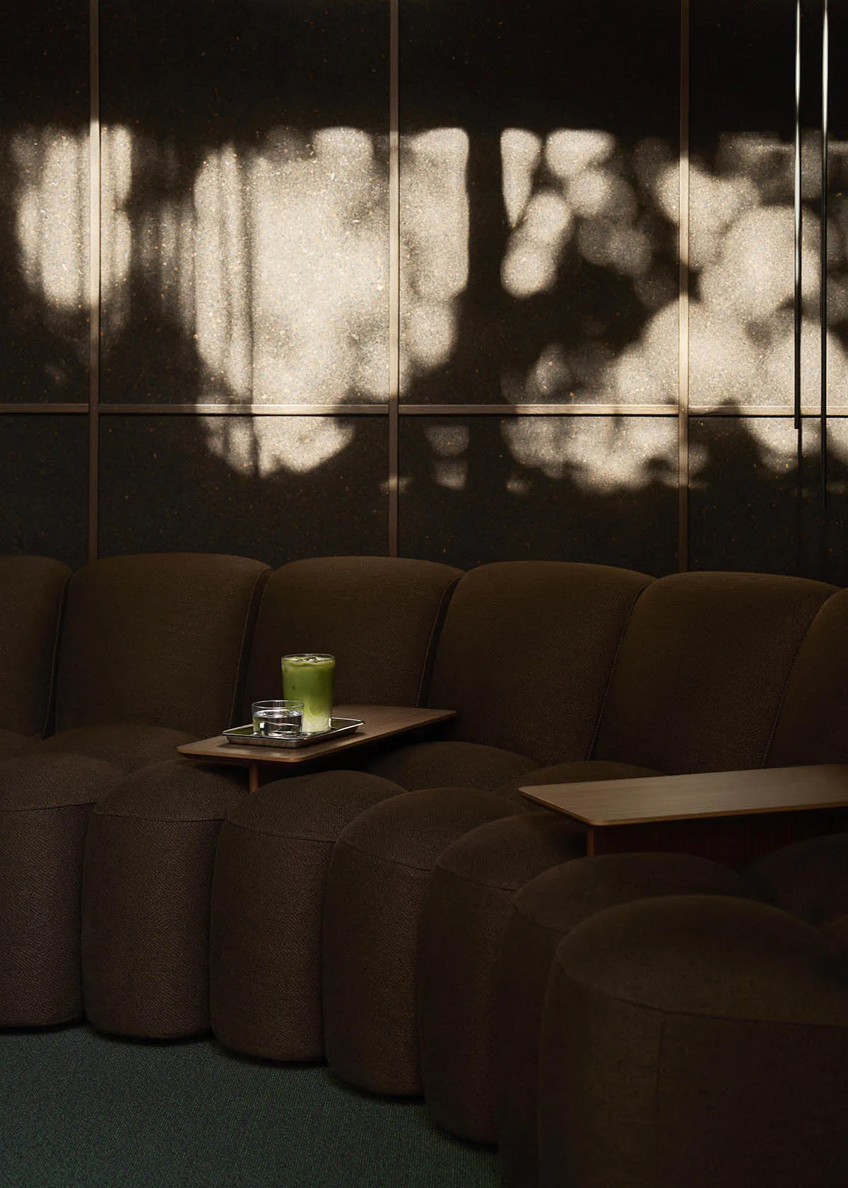 A curved brown boucle fabric modular sofa with a wooden side table holding a glass of iced matcha latte and a glass of water, set against a dark speckled wall with dramatic tree-like light and shadow patterns.