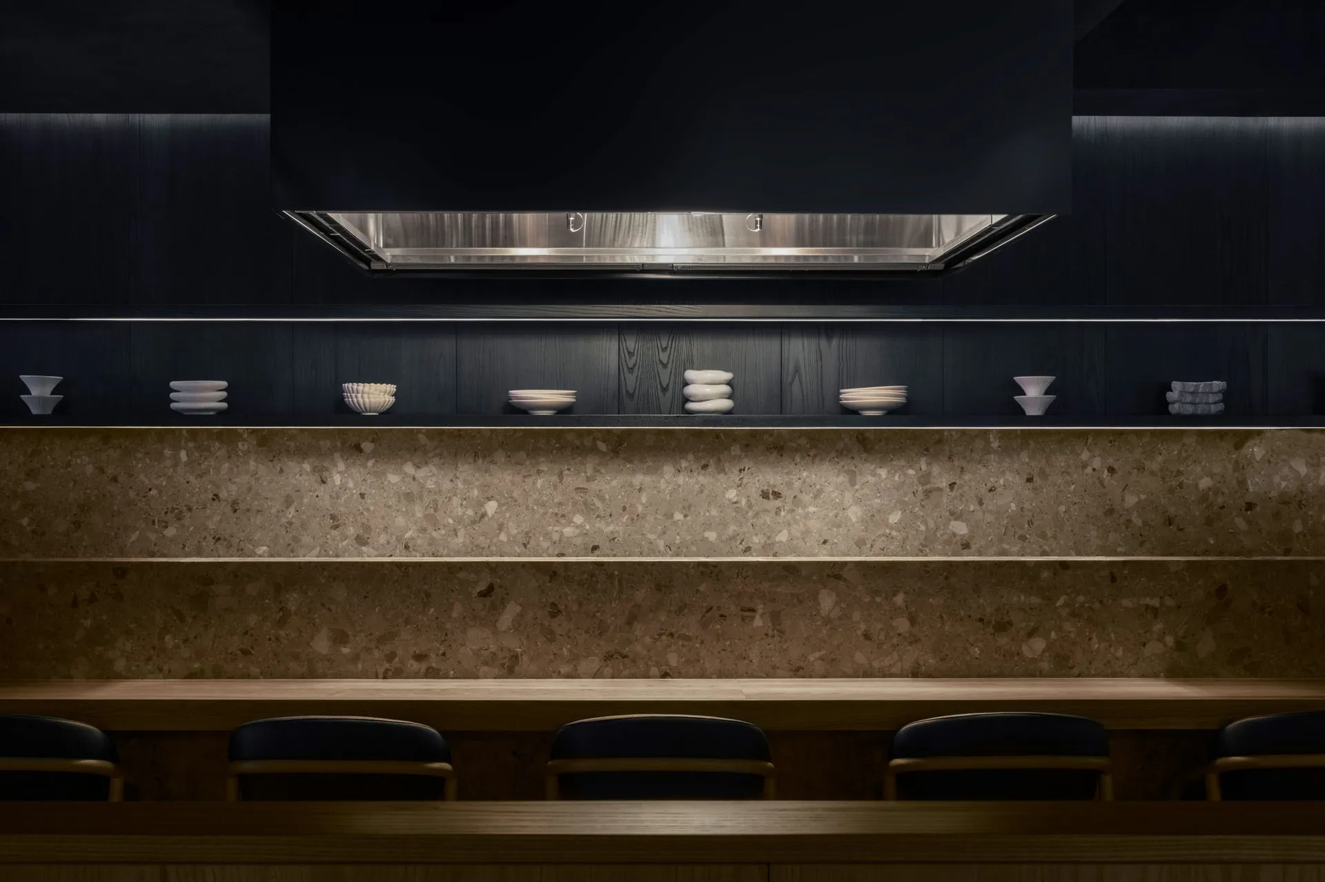 Modern dark kitchen featuring a long terrazzo countertop, light wood dining counter with navy blue chairs, open floating shelves displaying white ceramics, and dramatic under-cabinet lighting.