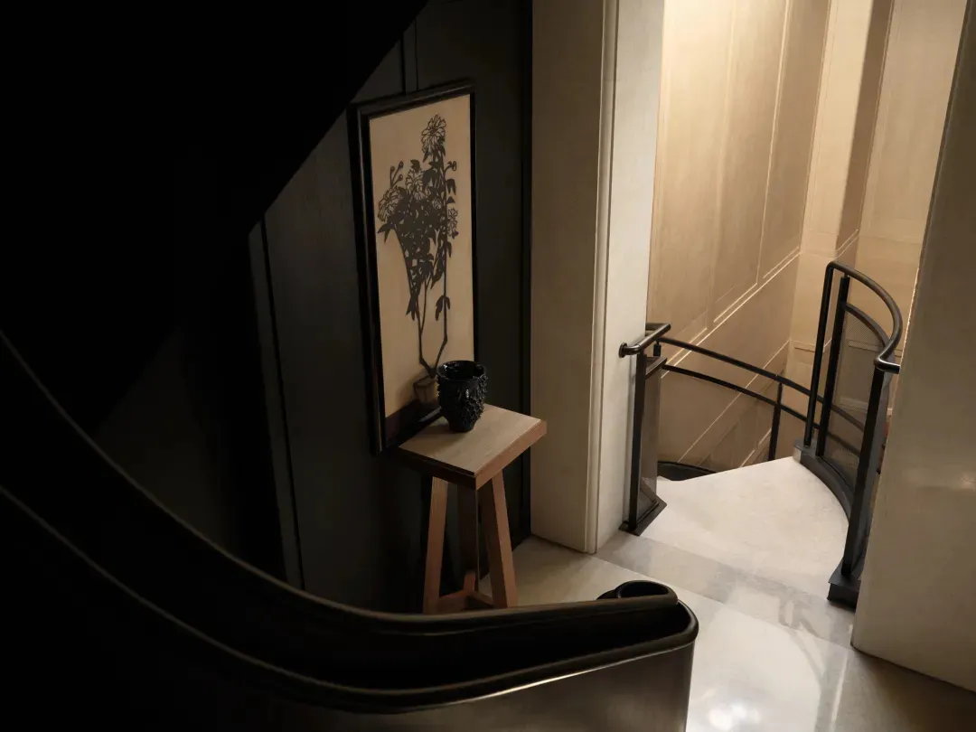 View of a modern curved staircase with black metal railing, light marble floor, oak console table, and large framed botanical wall art on a dark wall.