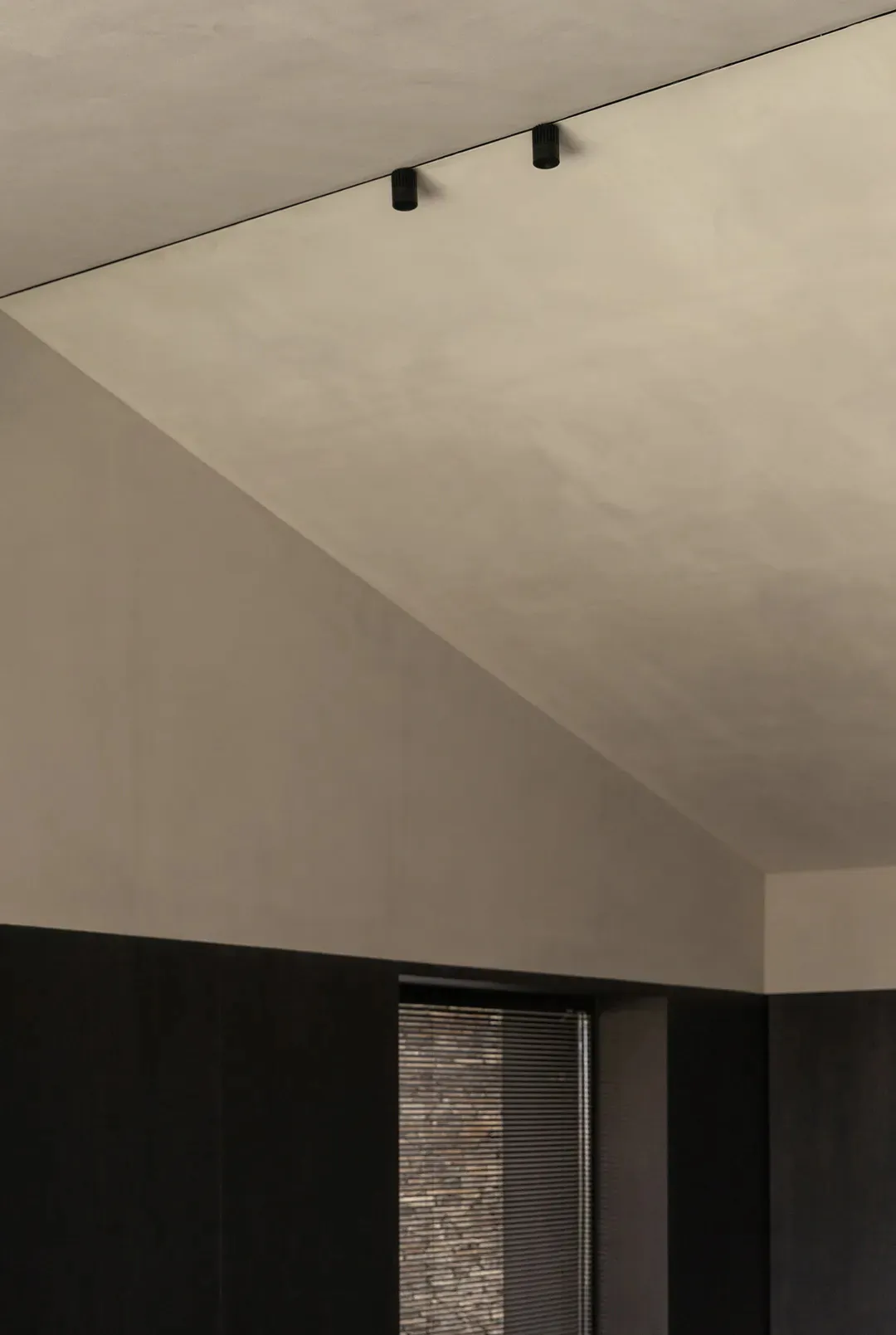View looking up at a raw concrete ceiling with two black cylindrical track lights mounted along a recessed line, above a dark wall opening with horizontal blinds.