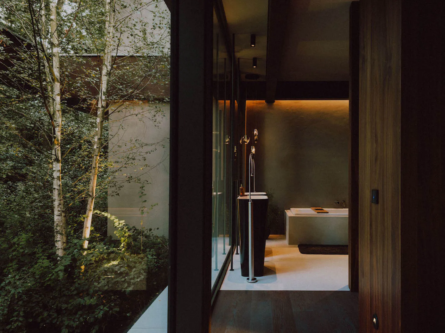 Modern minimalist bathroom interior featuring a freestanding black tub, chrome floor-mounted faucet, white rectangular bathtub, walnut wood door and paneling, textured concrete walls, and large glass windows overlooking a dense green forest with birch trees.