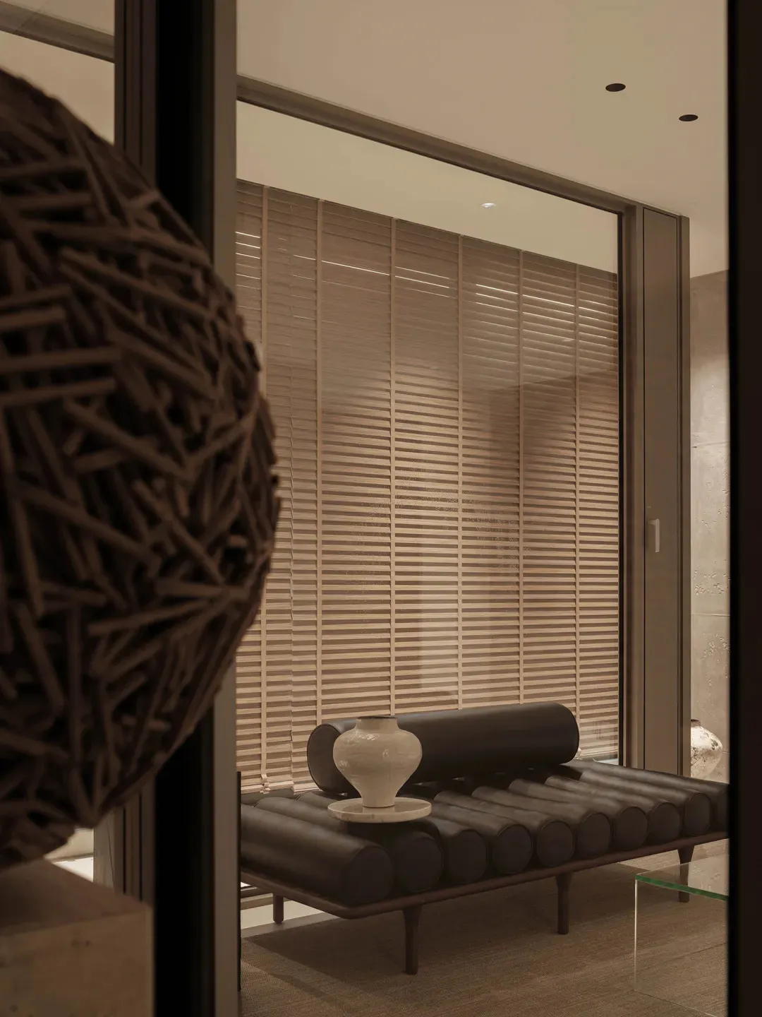 Modern minimalist living space featuring a dark leather tubular daybed with a white ceramic vase on a marble tray, set against large windows with horizontal wooden blinds, viewed through a dark sculptural art piece in the foreground.