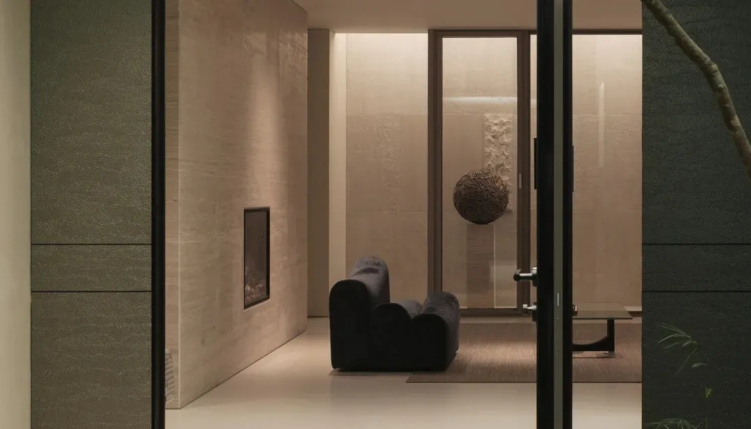 View into a modern minimalist living room featuring travertine walls, a sculptural black velvet sofa, a suspended woven spherical sculpture, and warm ambient lighting.