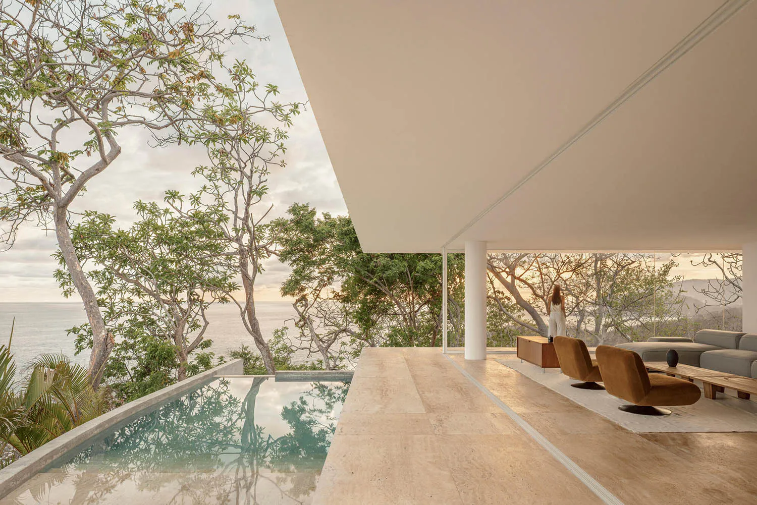 Modern open-air living room with travertine floors, brown velvet swivel chairs, a low wooden coffee table, and a long infinity pool overlooking the ocean and lush trees at sunset.