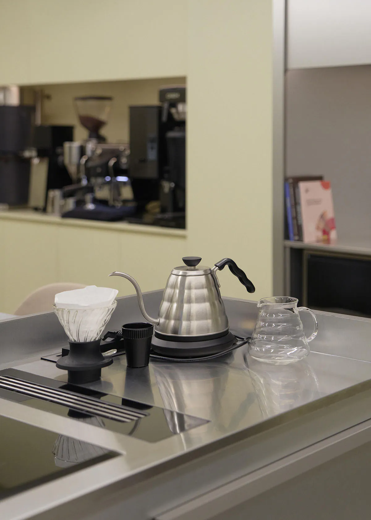 Stainless steel gooseneck kettle, glass pour-over dripper with paper filter, and glass server arranged on a reflective stainless steel countertop in a minimalist kitchen.