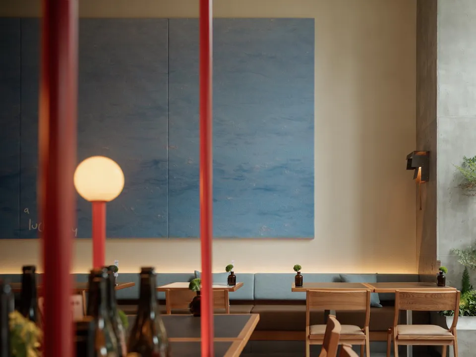 Modern restaurant interior featuring a large textured blue painting, built-in upholstered banquette seating, light oak wooden tables and chairs, and red structural poles in the foreground.