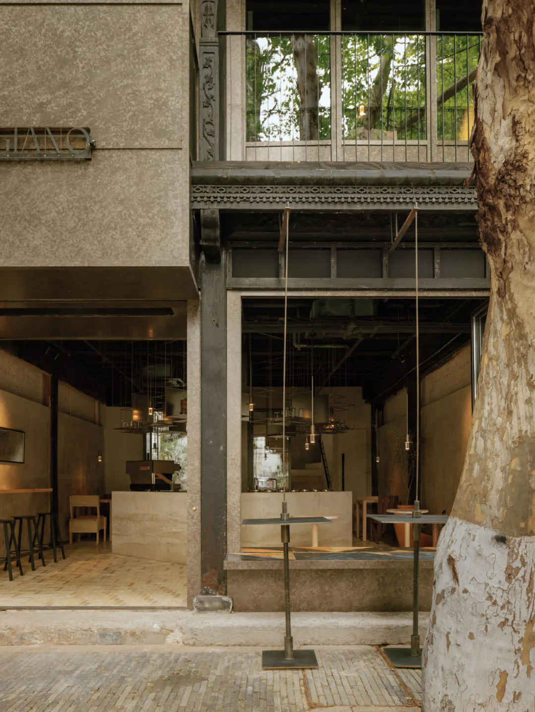 Exterior and interior view of a modern industrial-style cafe with concrete counters, high ceilings, hanging pendant lights, wooden herringbone flooring, and a large tree in the foreground.