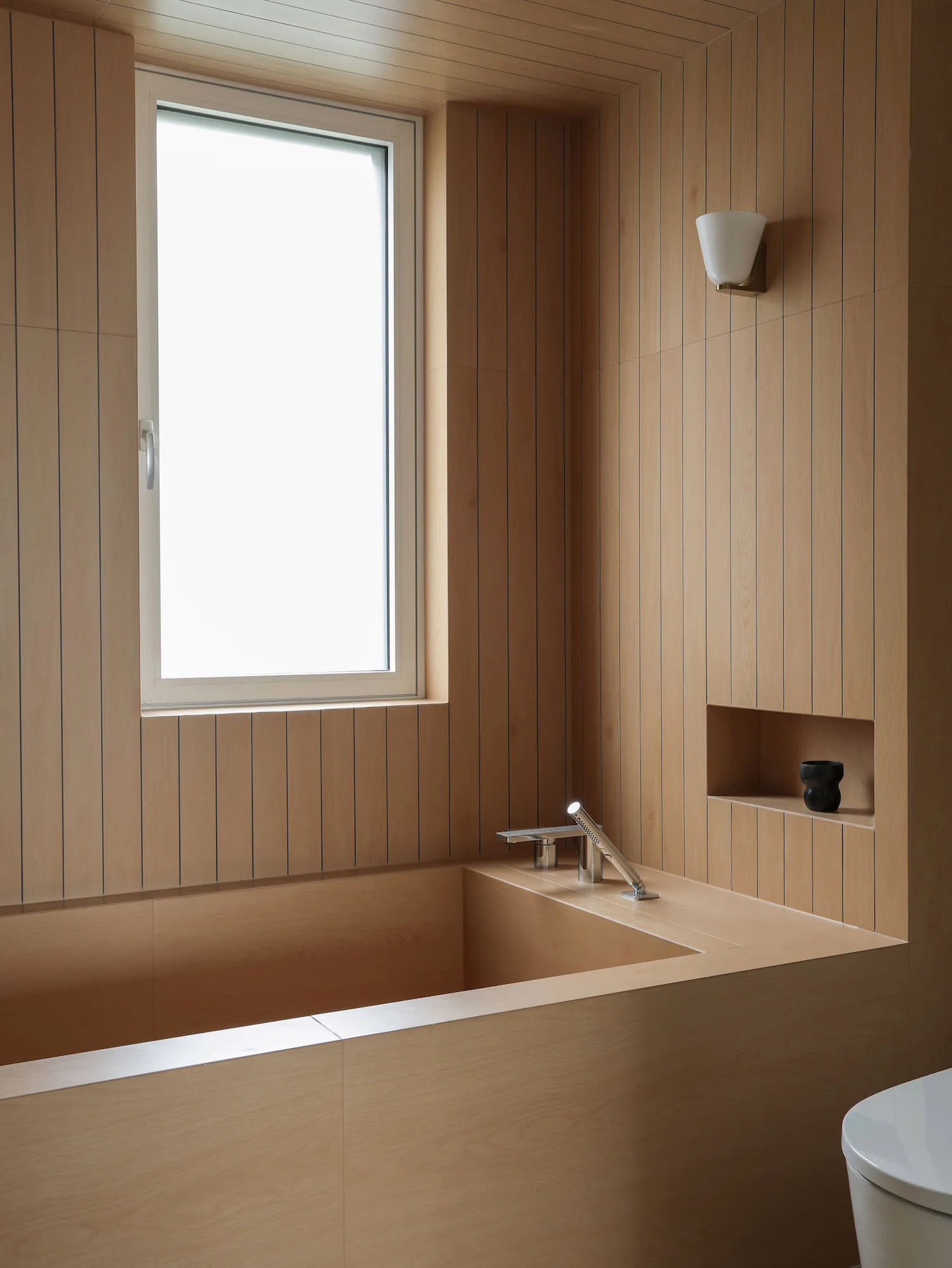 Modern minimalist bathroom featuring a built-in corner wooden bathtub, light oak vertical wall paneling, a large window, and a chrome wall-mounted faucet.