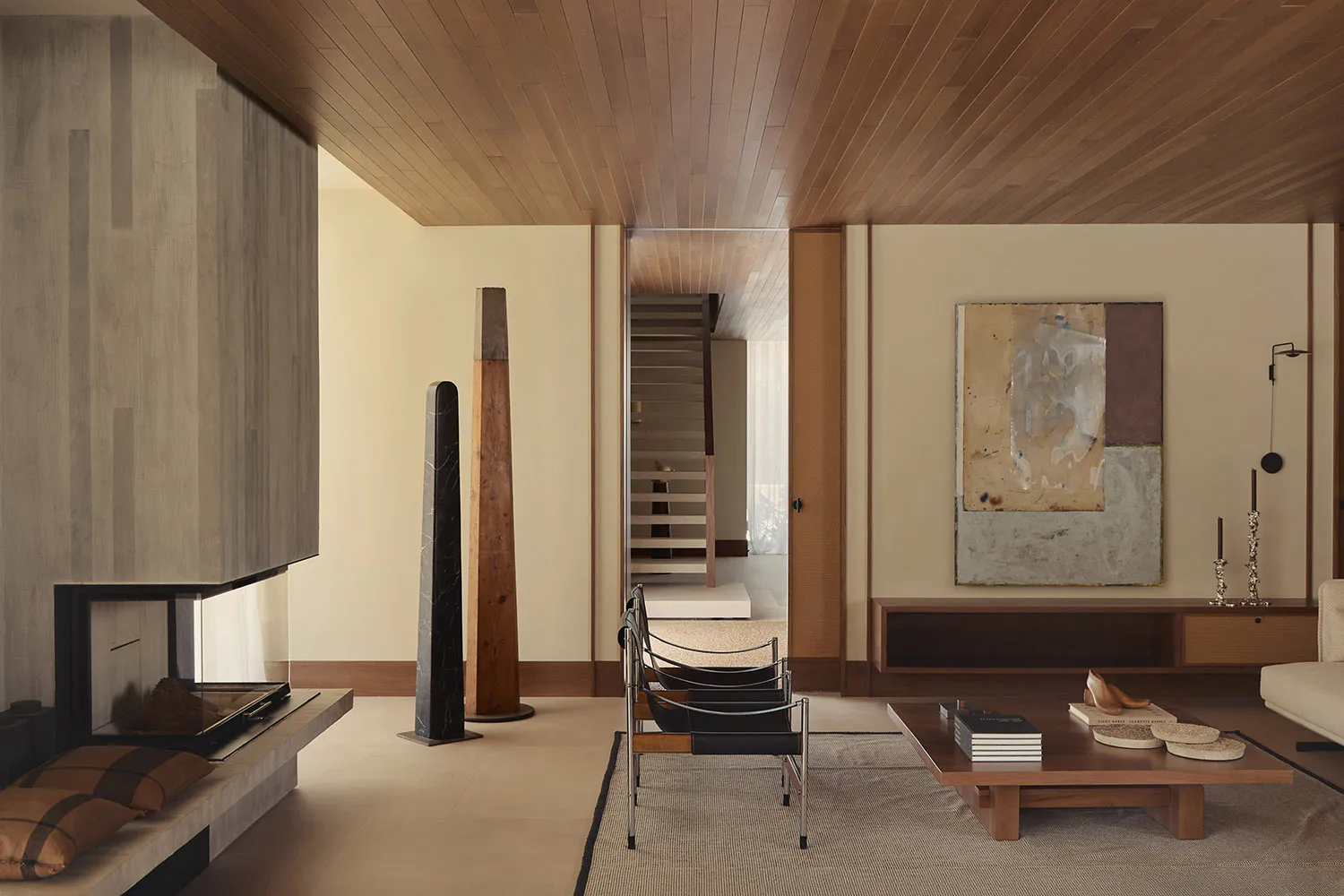 Modern minimalist living room with wood-paneled ceiling, floating fireplace, sculptural wooden and stone totems, black leather and chrome chairs, low wooden coffee table, and large abstract painting on a cream wall.