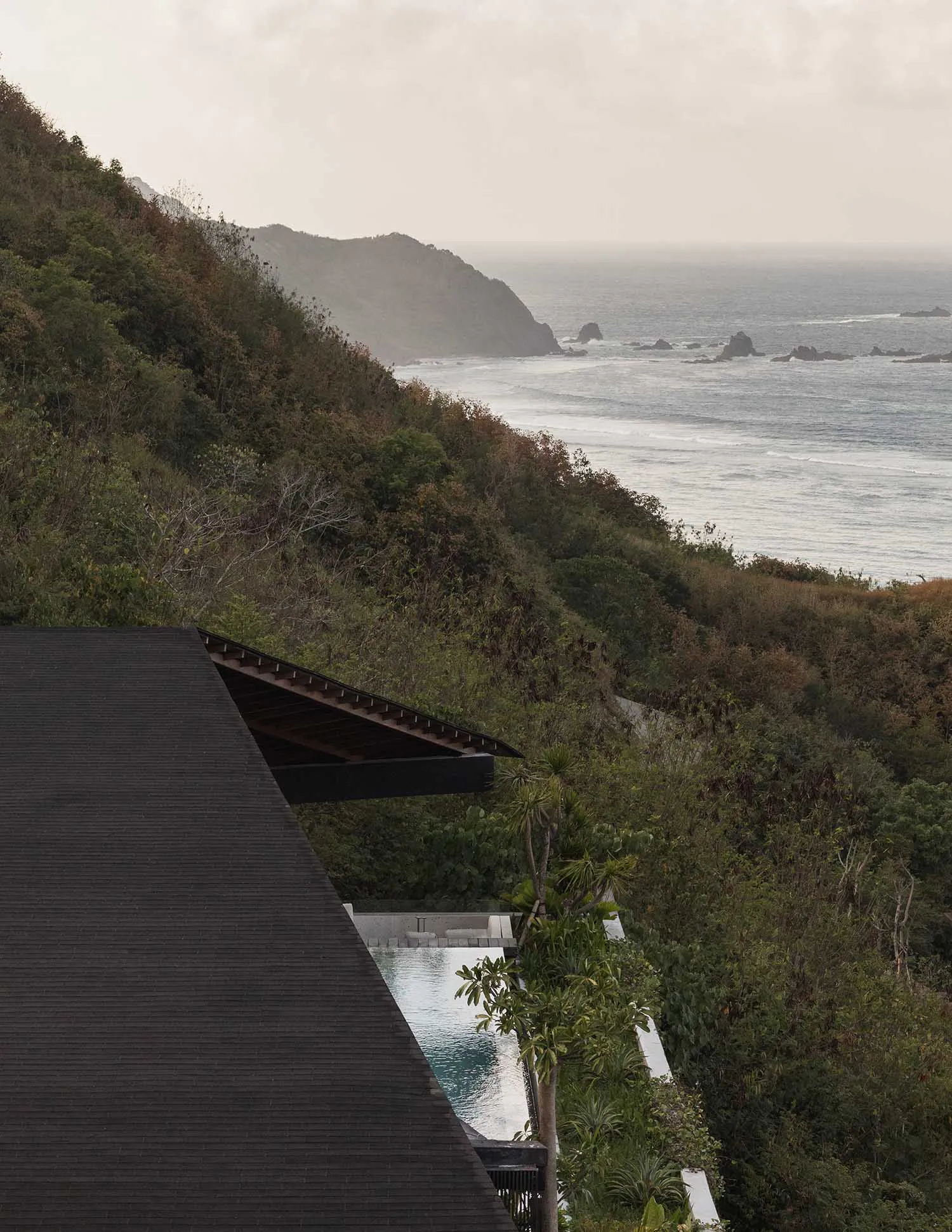 View from above of a modern villa with a dark timber roof and an infinity pool perched on a lush green cliffside overlooking the ocean and rocky coastline.
