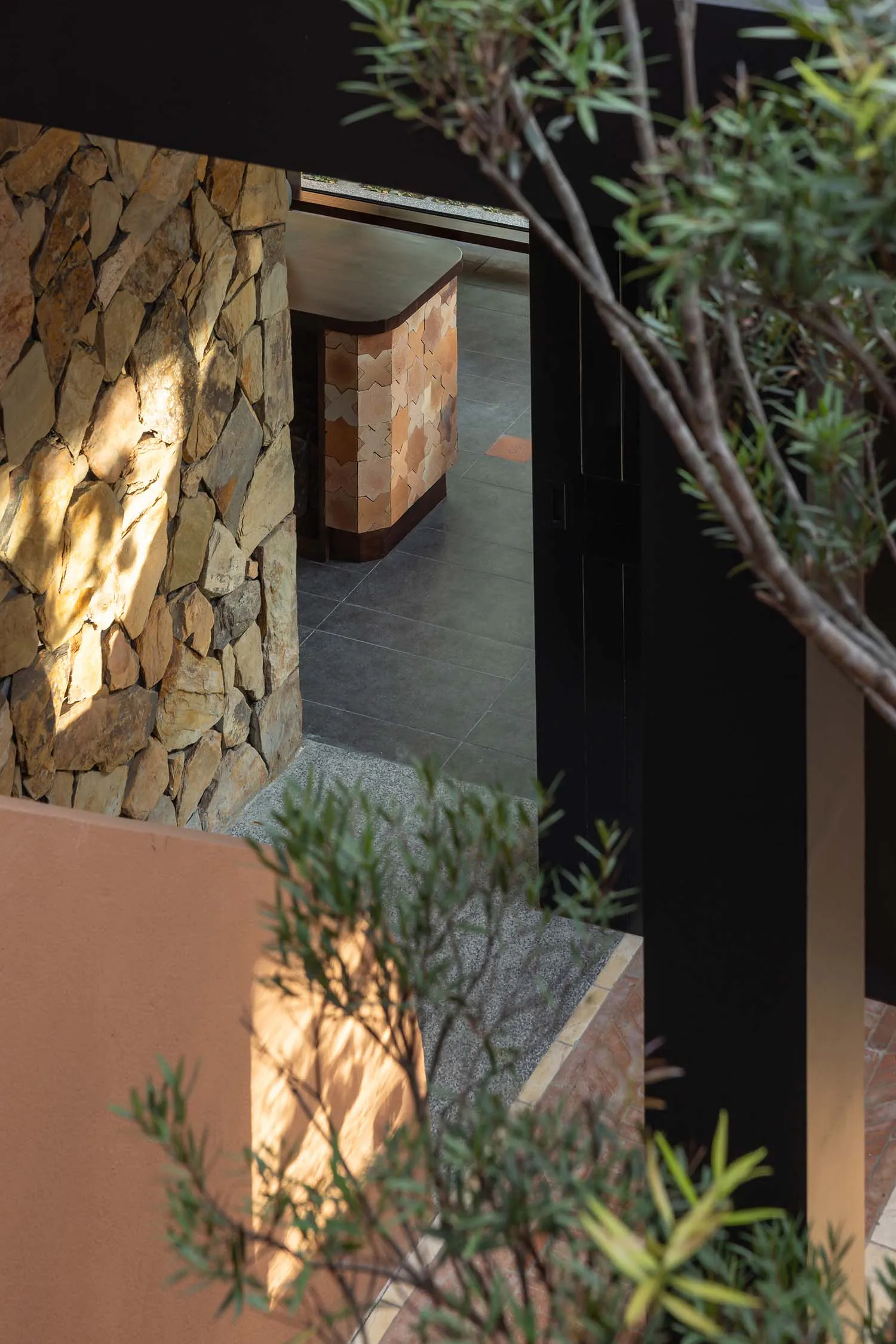 View through foliage of a sunlit natural stone wall and a modern entryway featuring terracotta patterned tiles and dark wood accents.