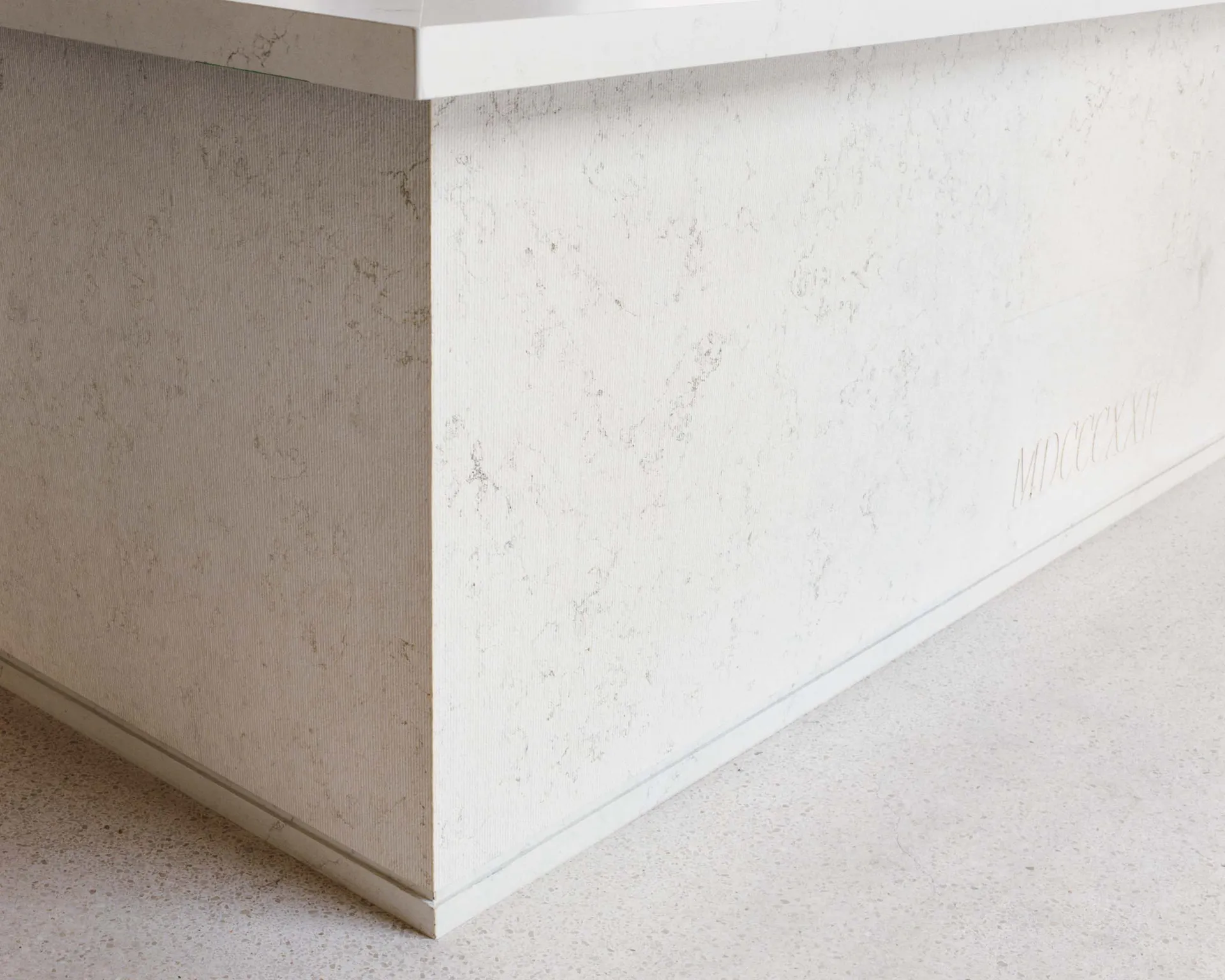 Close-up view of the corner of a white concrete kitchen island with visible aggregate texture and the Roman numerals MDCXXXII engraved on the side panel, set on a terrazzo floor.