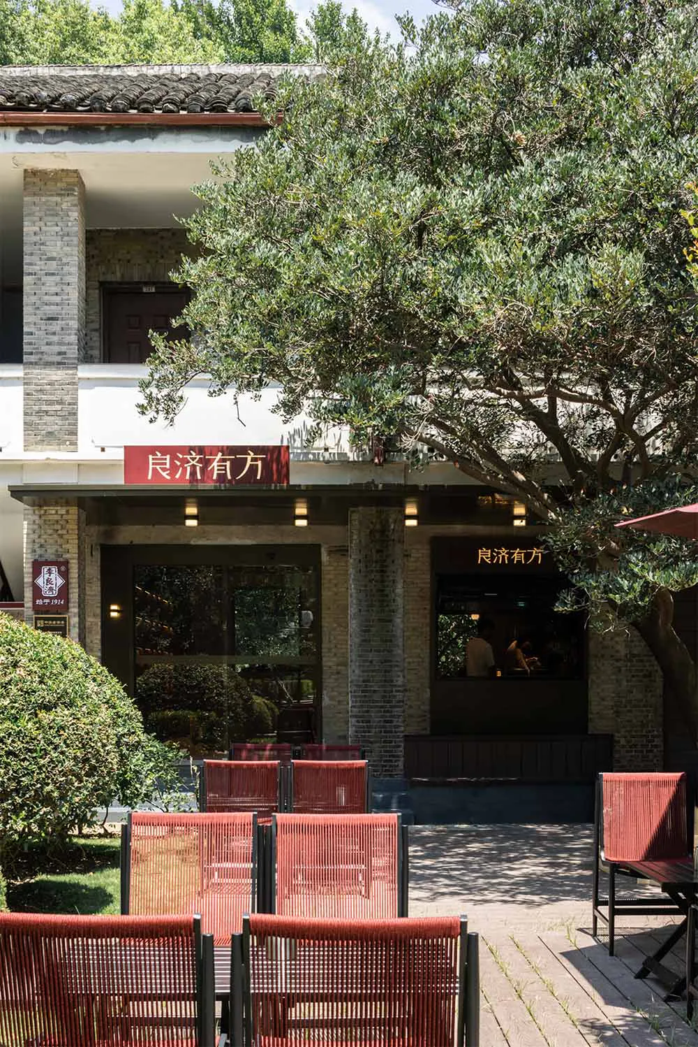 Outdoor seating area of a heritage building with red rope-weave chairs on a wooden deck, surrounded by lush greenery and a large tree, with Chinese signage reading 良濟有方.