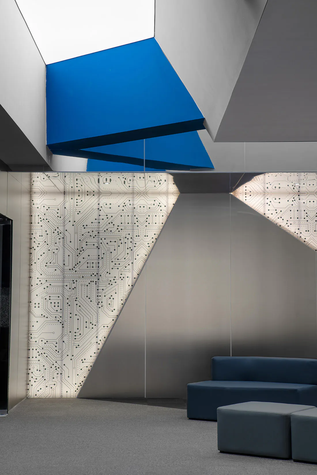 Modern corporate lobby interior featuring a large backlit wall with white circuit board patterns, angular blue ceiling panels, and blue modular seating on grey carpet.