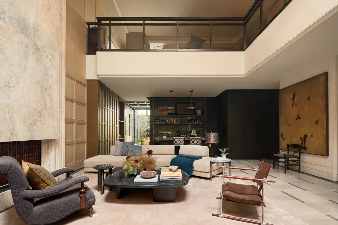Spacious double-height modern luxury living room with marble walls, large sectional sofa, teal throw, dark wood armchair, and upper mezzanine level.