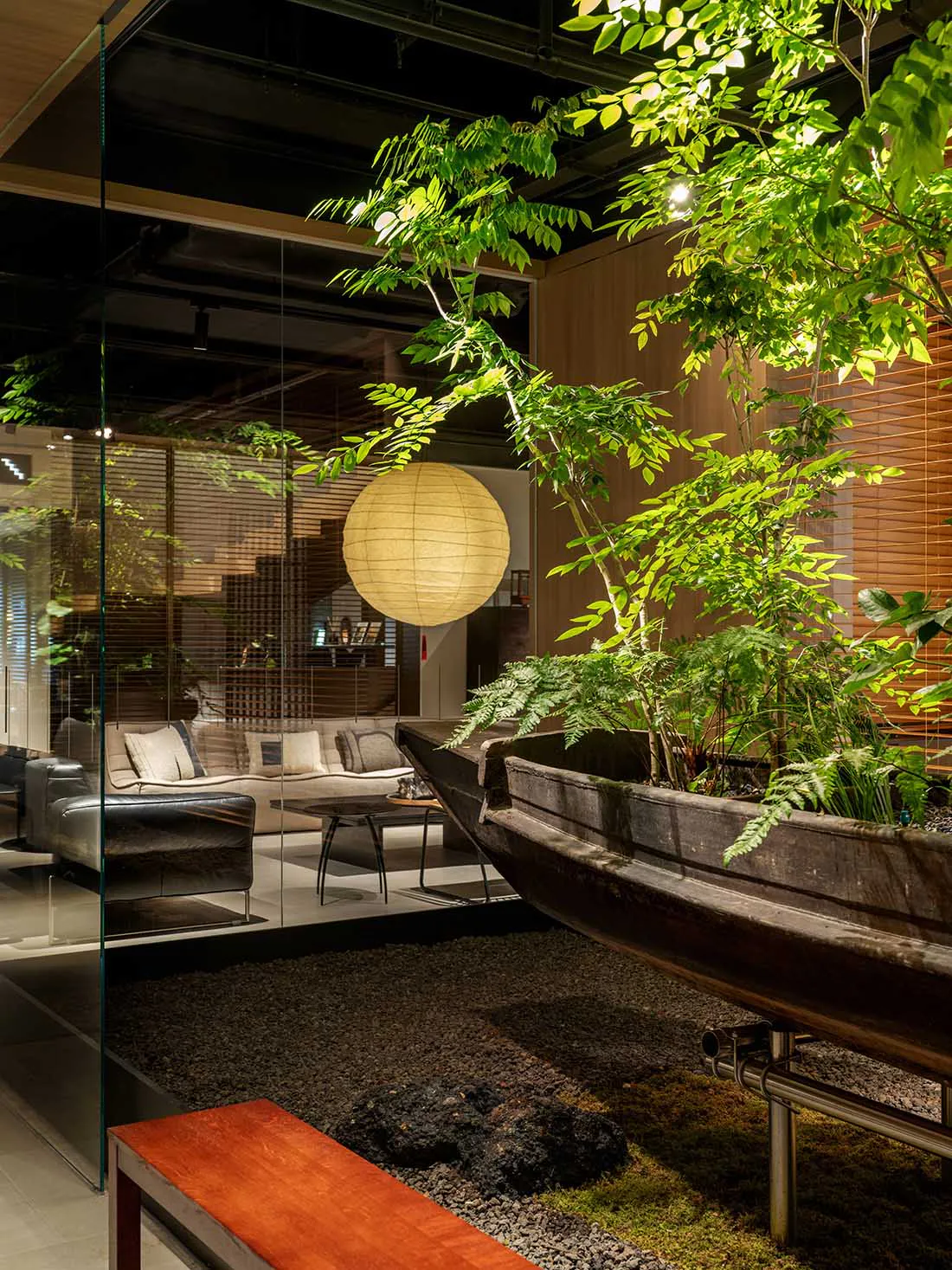 Modern living room viewed through glass with a large repurposed wooden boat used as a planter filled with ferns and small trees, paper lantern pendant light, dark leather armchair, and gravel floor in an indoor garden setting.
