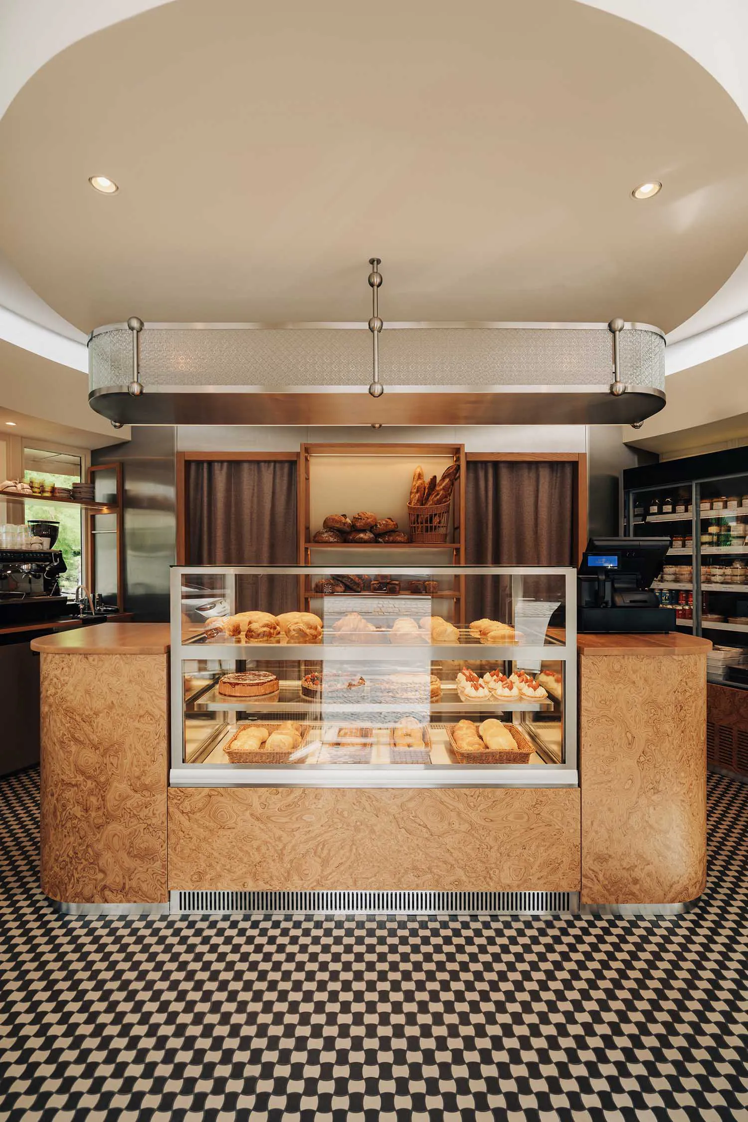 Modern bakery interior with a curved counter finished in burl wood, a large glass pastry display case filled with croissants and cakes, black and white checkered floor, and a large circular ceiling detail.