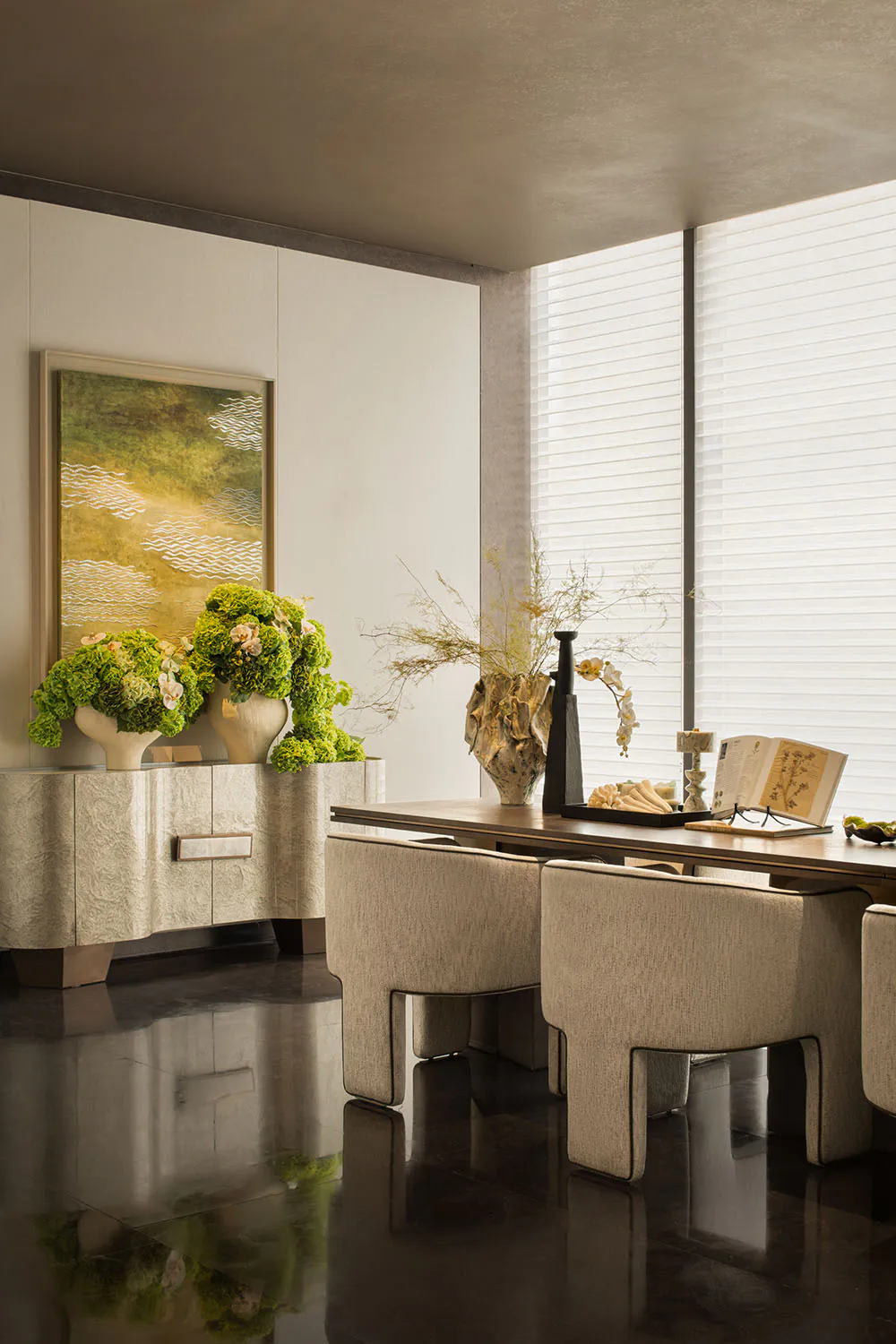 Modern minimalist dining room featuring a textured beige sideboard with large green hydrangea arrangements, a wooden dining table with sculptural dried floral centerpiece, beige upholstered dining chairs, and a large abstract landscape painting on the wall.