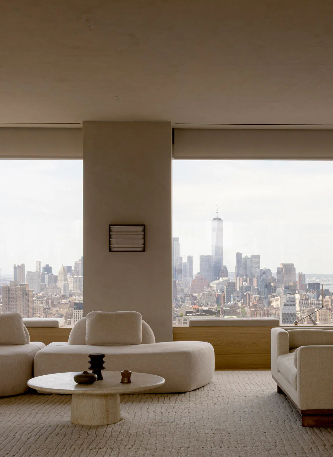 Modern minimalist living room with curved beige sofas, a round travertine coffee table, and floor-to-ceiling windows overlooking the New York City skyline including One World Trade Center.