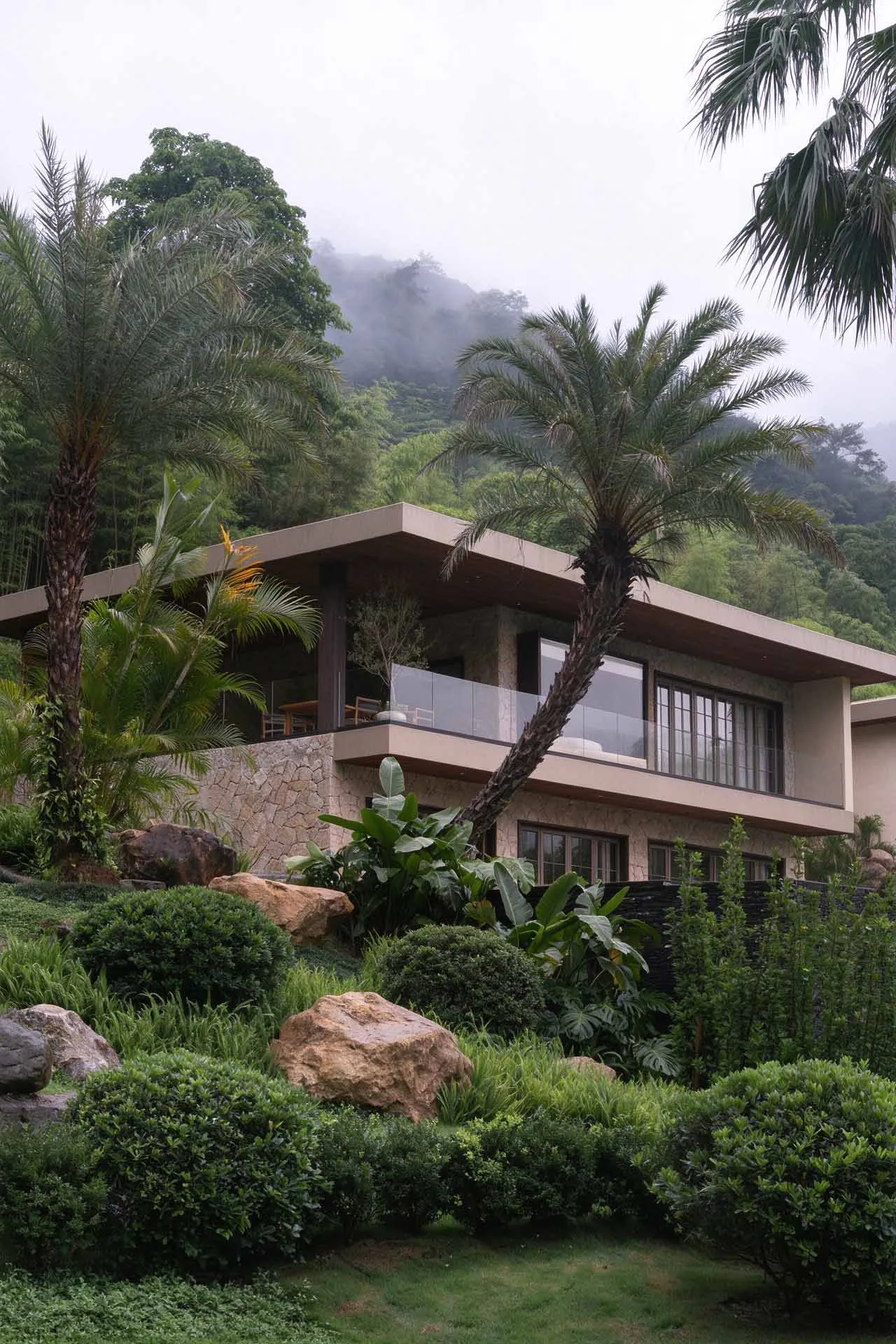 Modern two-story concrete and stone villa with large glass windows and balconies, surrounded by palm trees, large boulders, manicured hedges and tropical plants on a misty hillside.