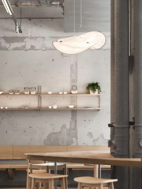 Interior view of a modern industrial-style cafe with exposed concrete walls, wooden tables and stools, floating wooden shelves displaying ceramics and plants, and a large sculptural white pendant lamp hanging from the ceiling.