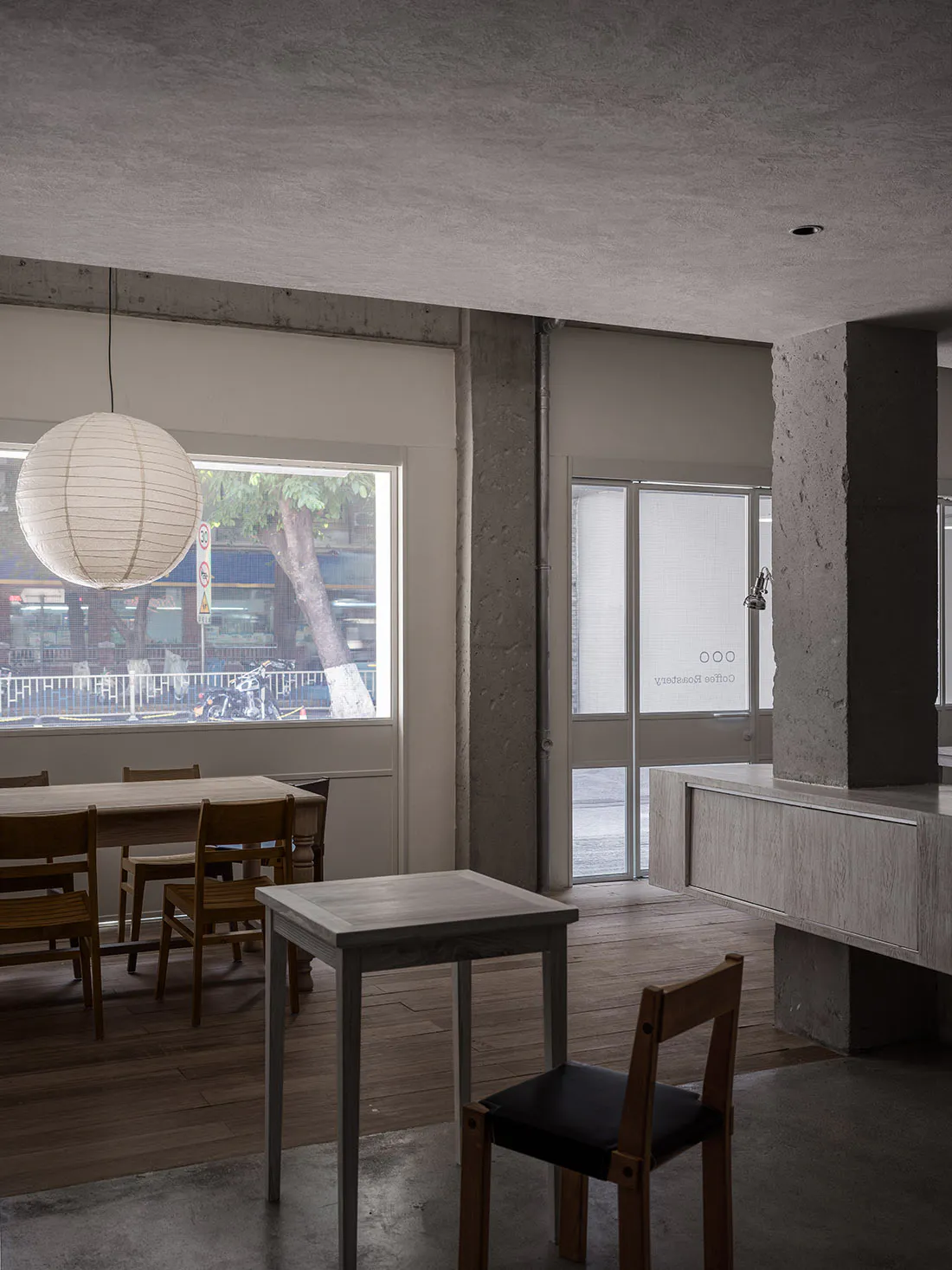 Interior view of a minimalist cafe featuring raw concrete walls and pillars, light wood flooring, wooden dining tables and chairs, a large white paper lantern, and a view of the street through large windows.