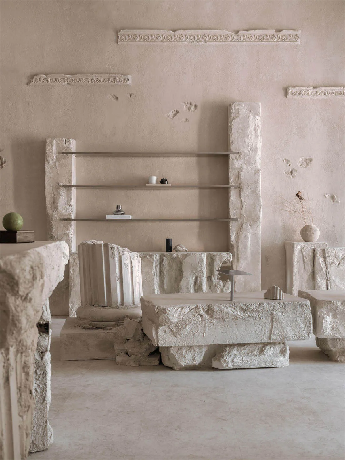 Minimalist living room featuring massive rough-hewn limestone blocks used as structural elements, coffee tables, and pedestals, with floating metal shelves mounted within a stone wall, decorative crown moldings on the upper wall, and neutral-toned accessories including a green sphere, dried botanicals, and sculptural objects.