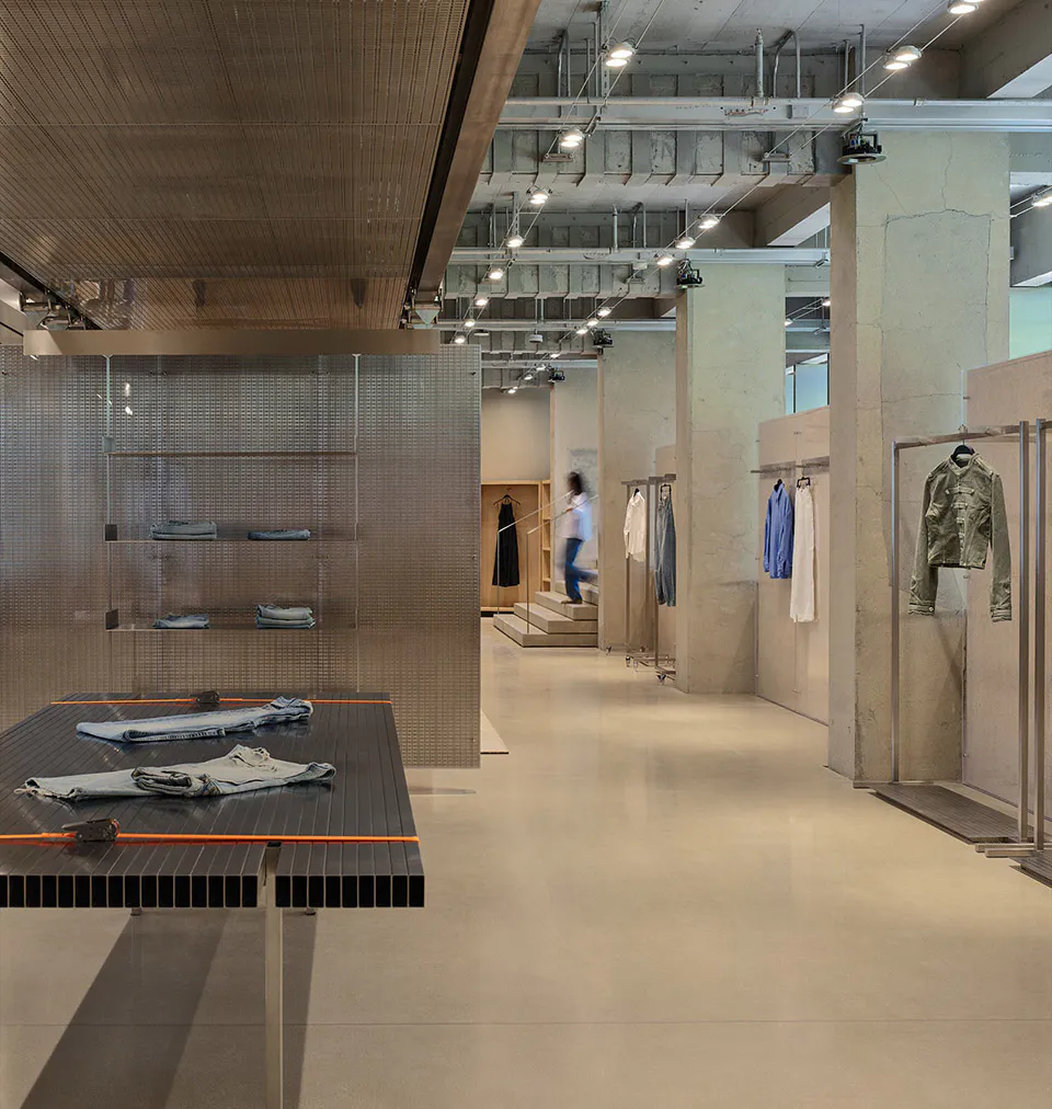 Modern industrial-style clothing boutique interior with raw concrete walls, polished concrete floor, metal mesh display shelves, and dark slatted display tables holding folded denim garments.