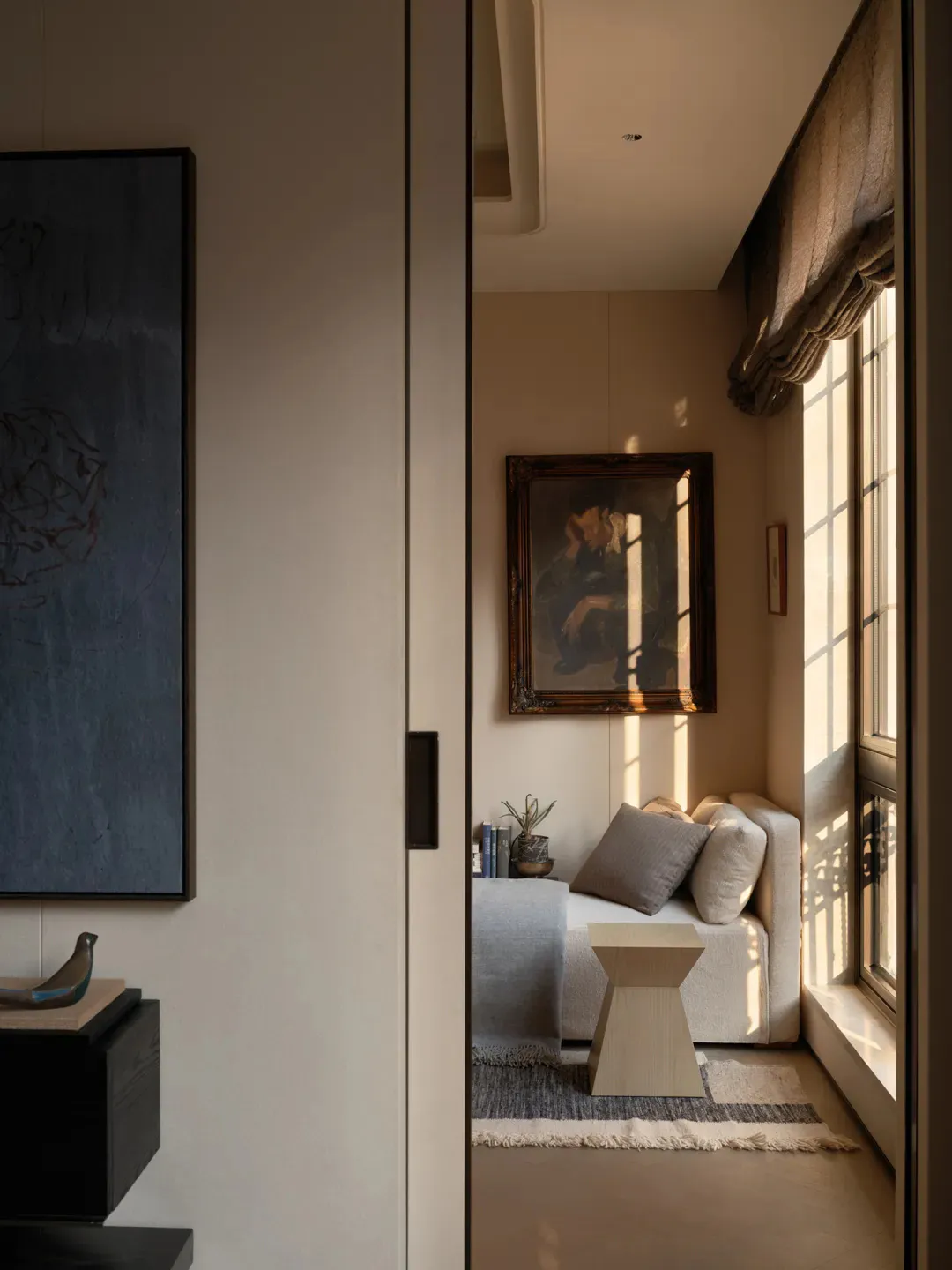 Minimalist reading nook featuring a built-in linen window seat, wooden side table, framed painting, and soft natural light from a large window with roman shade.